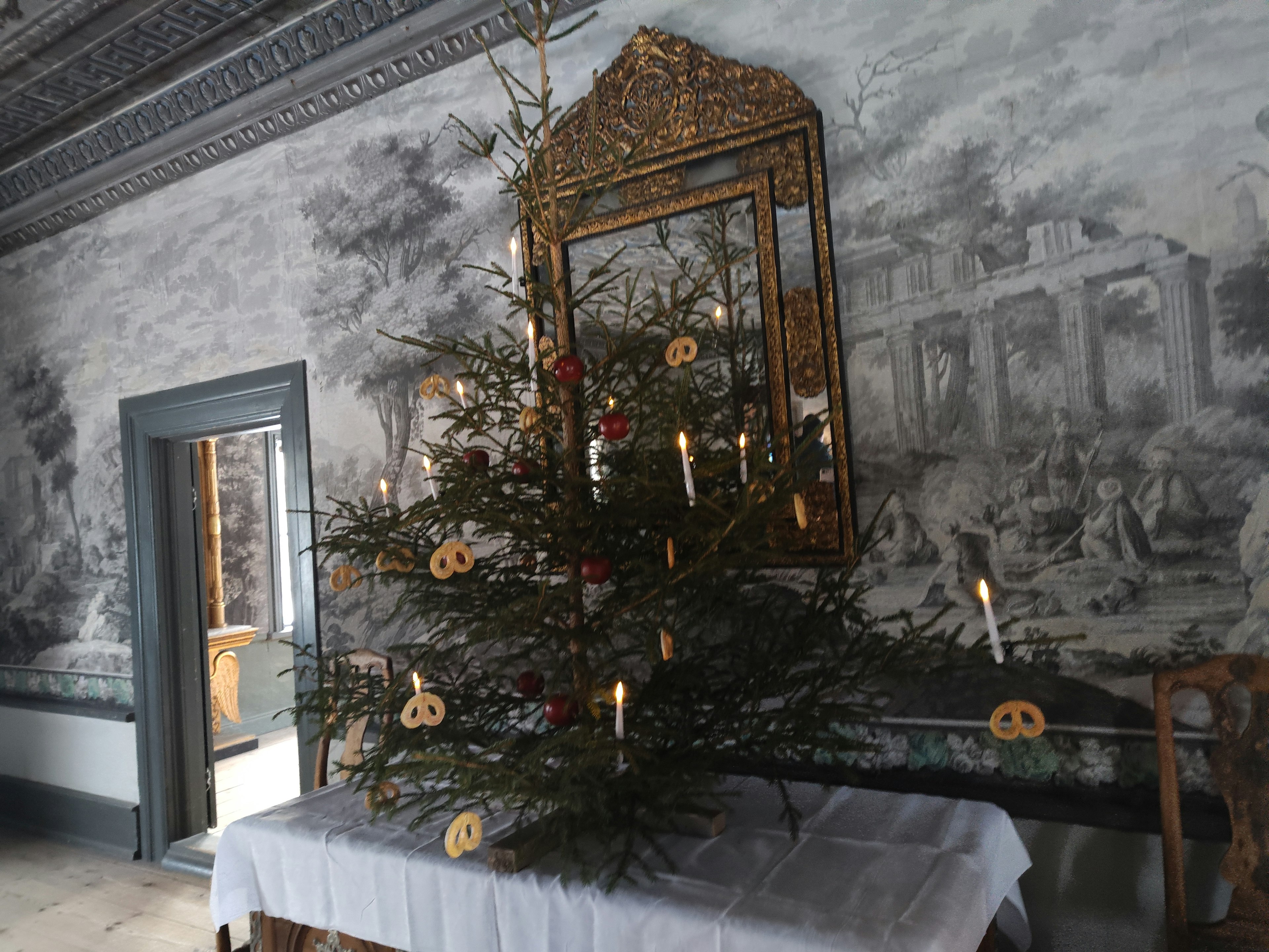 A decorated Christmas tree with candles and ornaments stands in an ornate room featuring vintage wall paintings