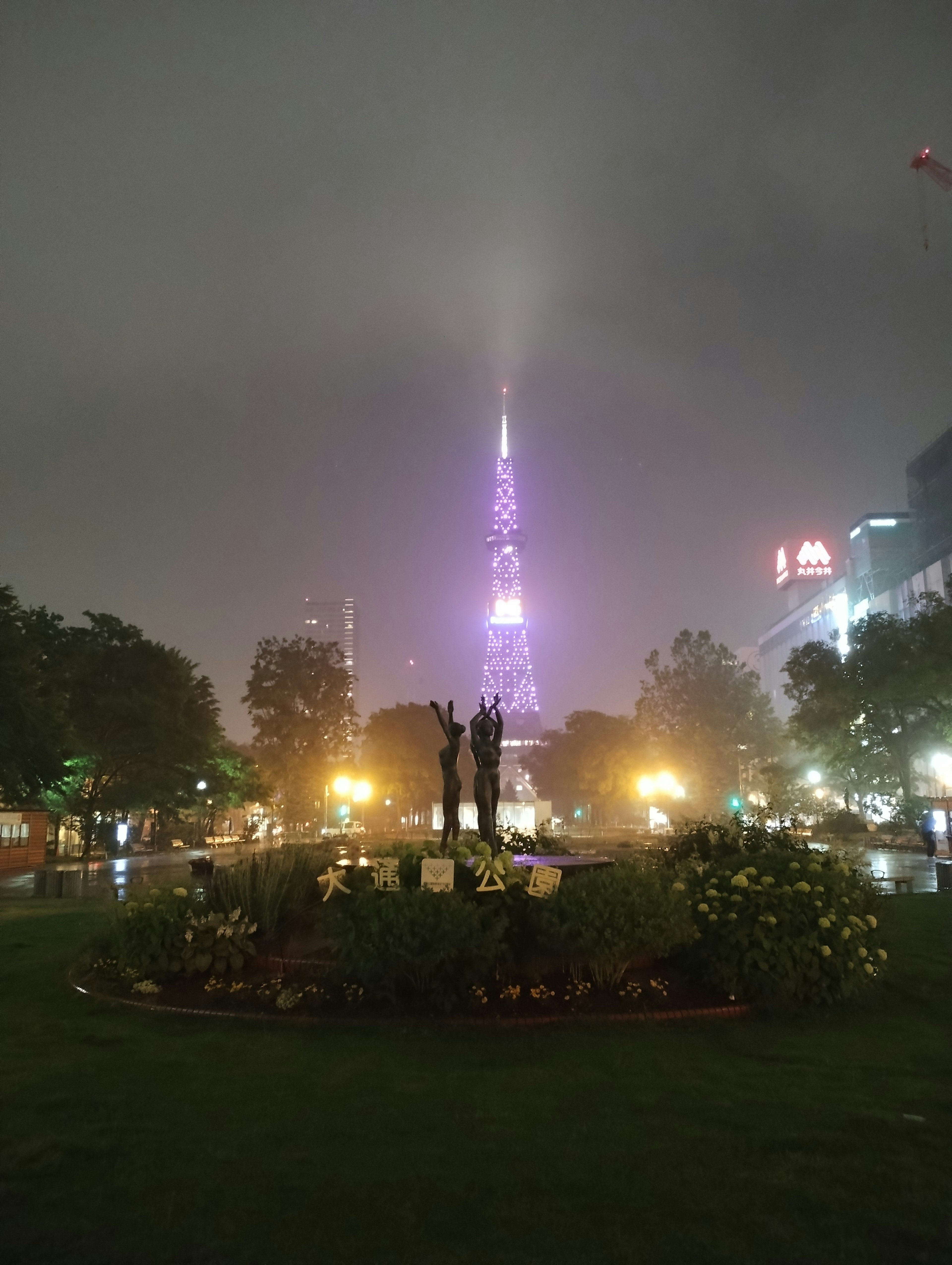 Estatua en un parque nocturno con una torre morada brillando en la niebla