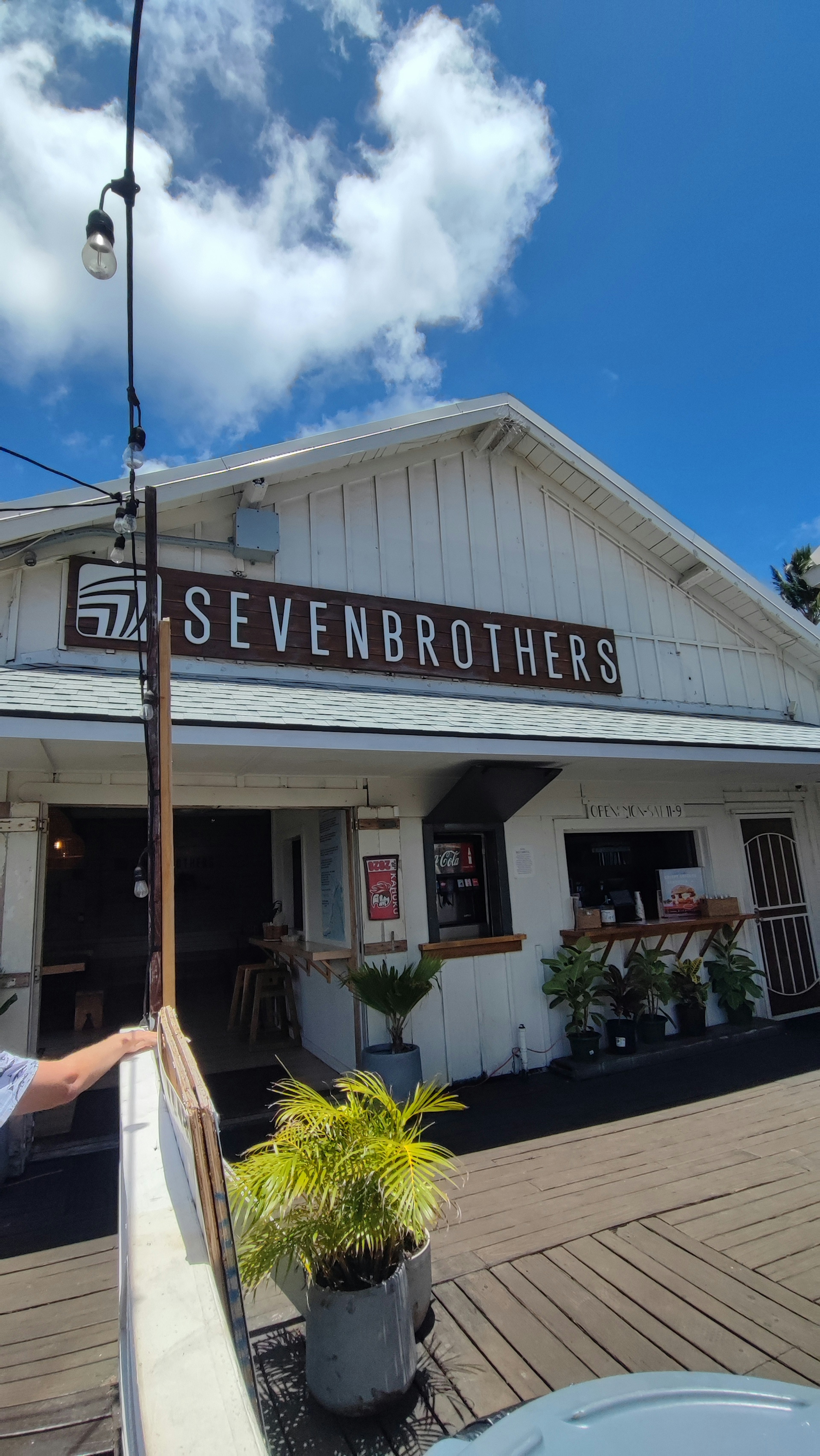 Exterior del restaurante Seven Brothers con cielo azul y nubes blancas y decoraciones de plantas