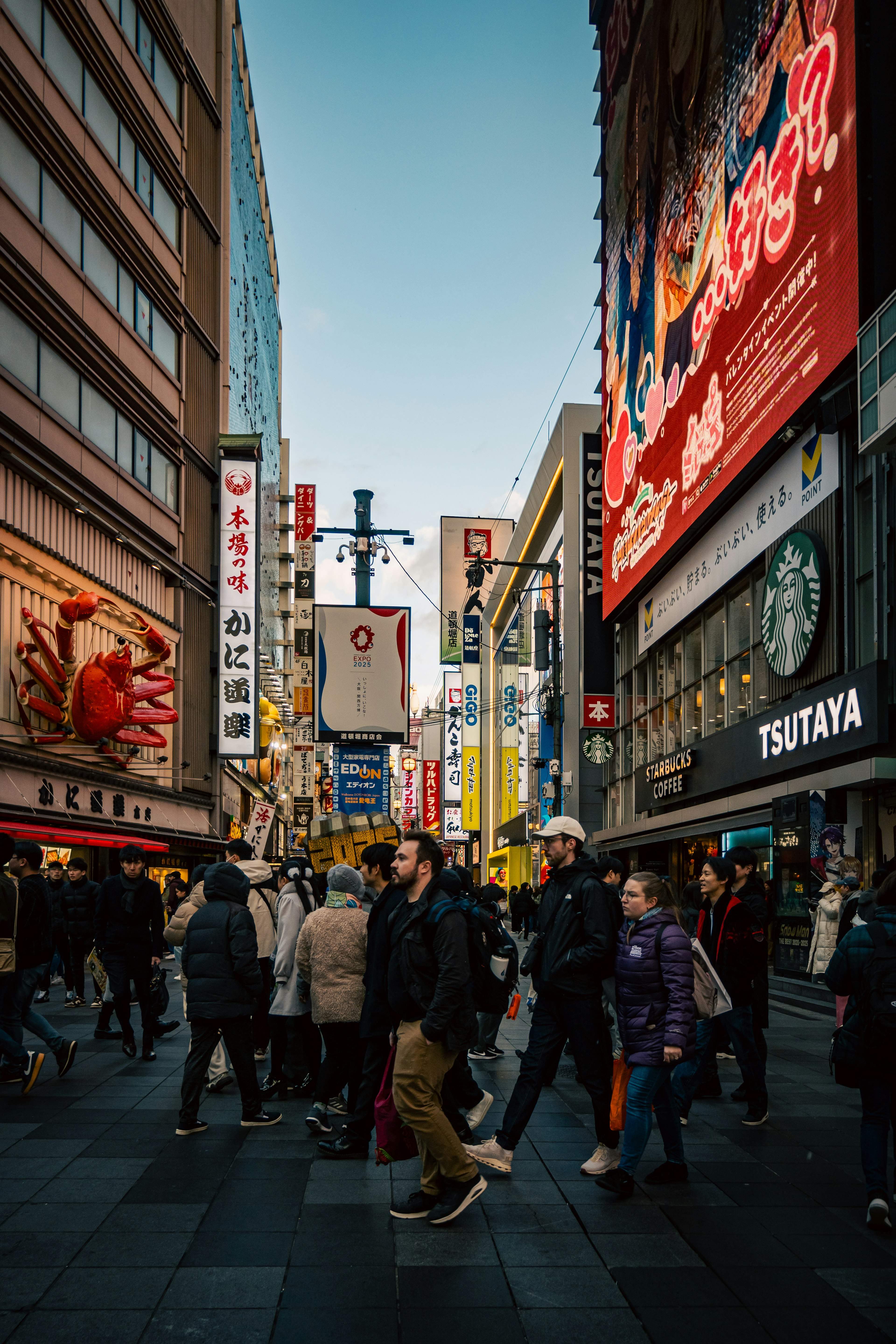 賑やかな街の風景で、多くの人々が交差点を渡る様子が描かれている