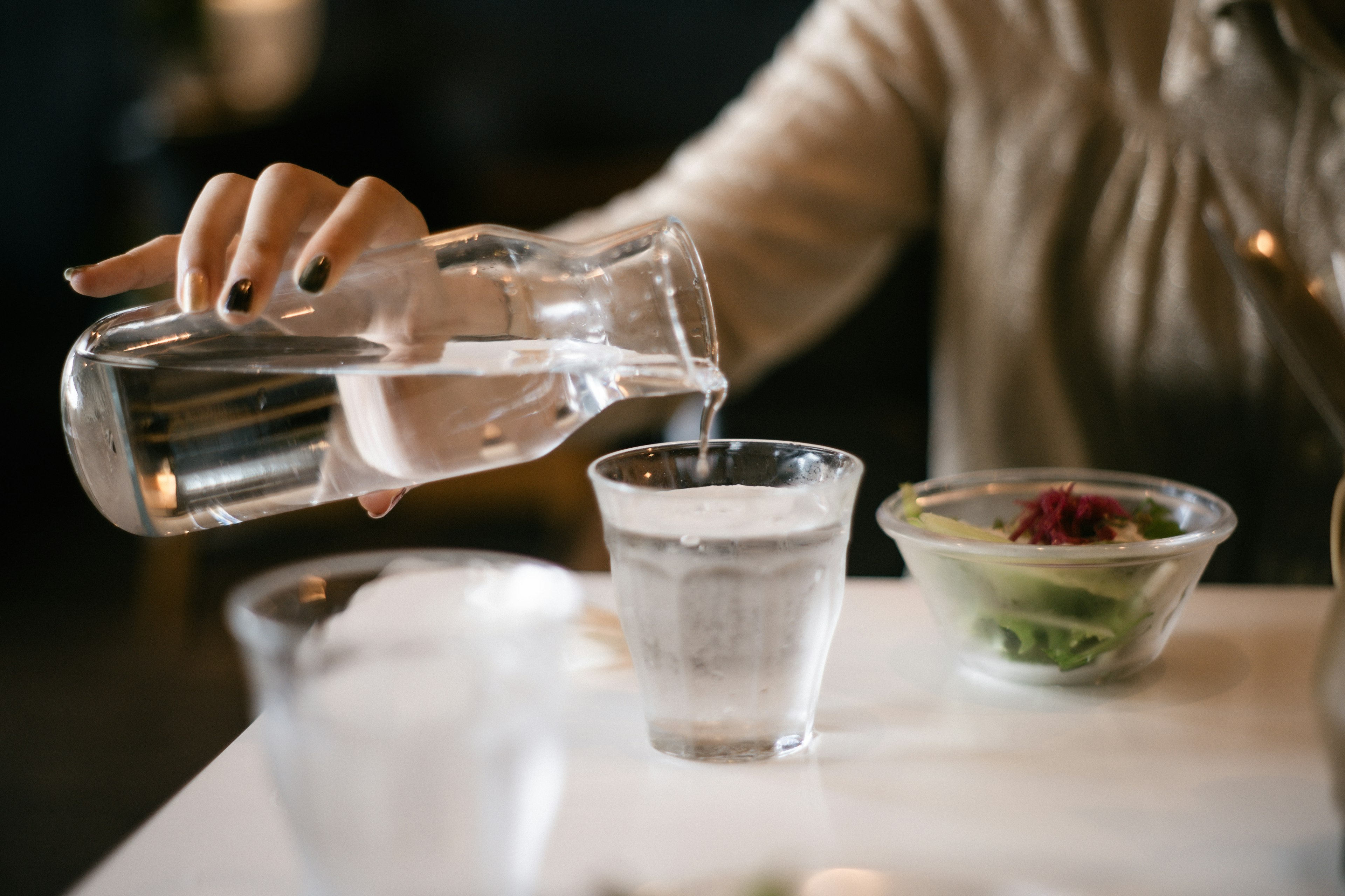 Sebuah tangan menuangkan air dari carafe kaca ke dalam gelas transparan di atas meja dengan salad