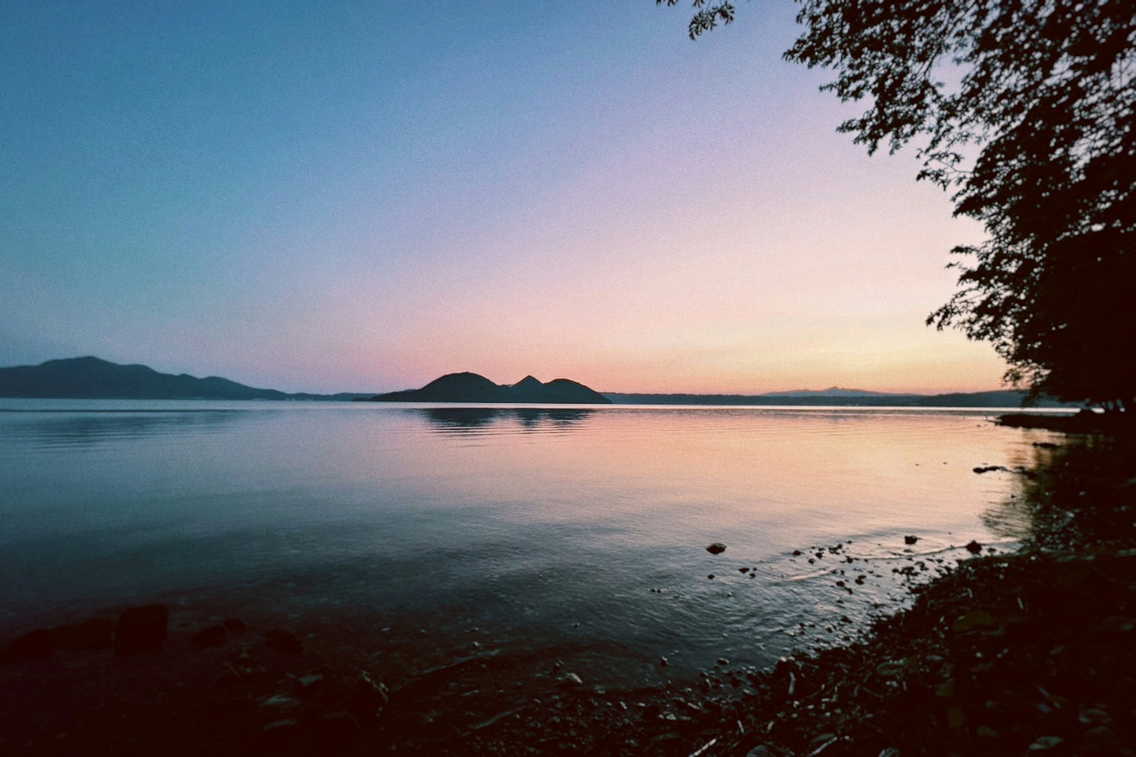 Danau tenang saat matahari terbenam dengan pegunungan yang terpantul di air dan gradasi warna