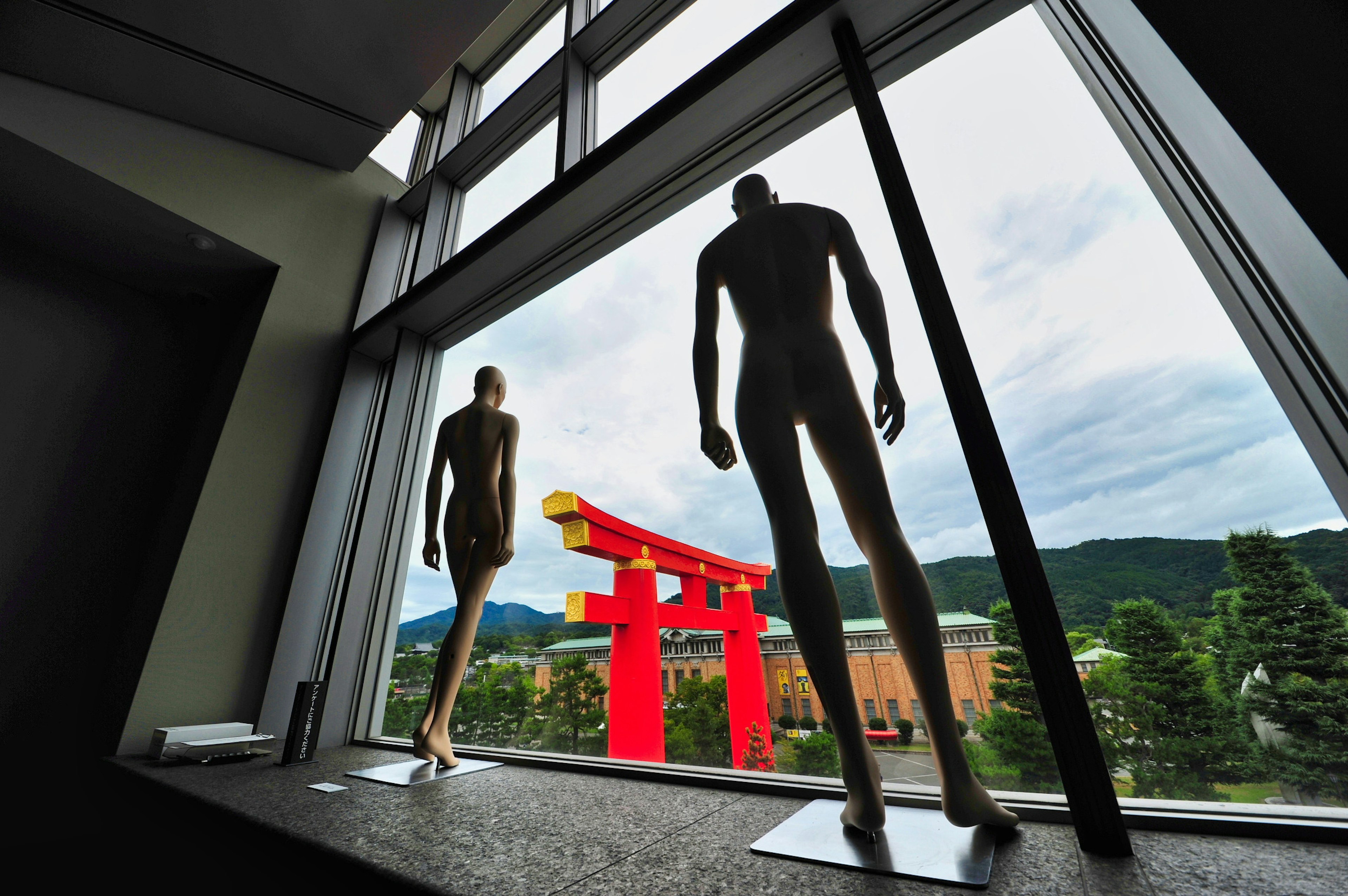 Silhouetten von zwei Schaufensterpuppen vor einem roten Torii, sichtbar durch das Fenster