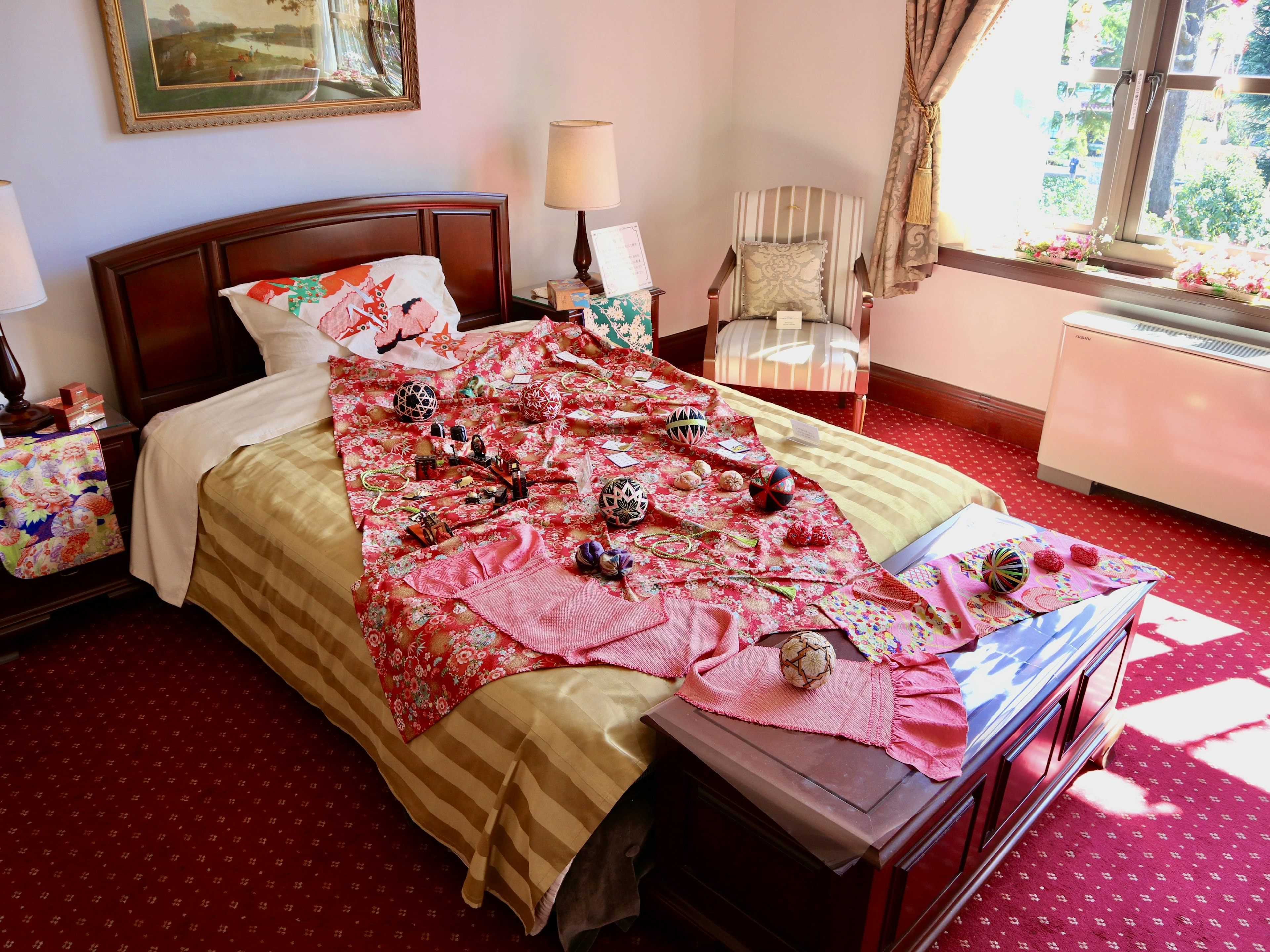 A room with a neatly made single bed covered in a pink blanket surrounded by scattered objects