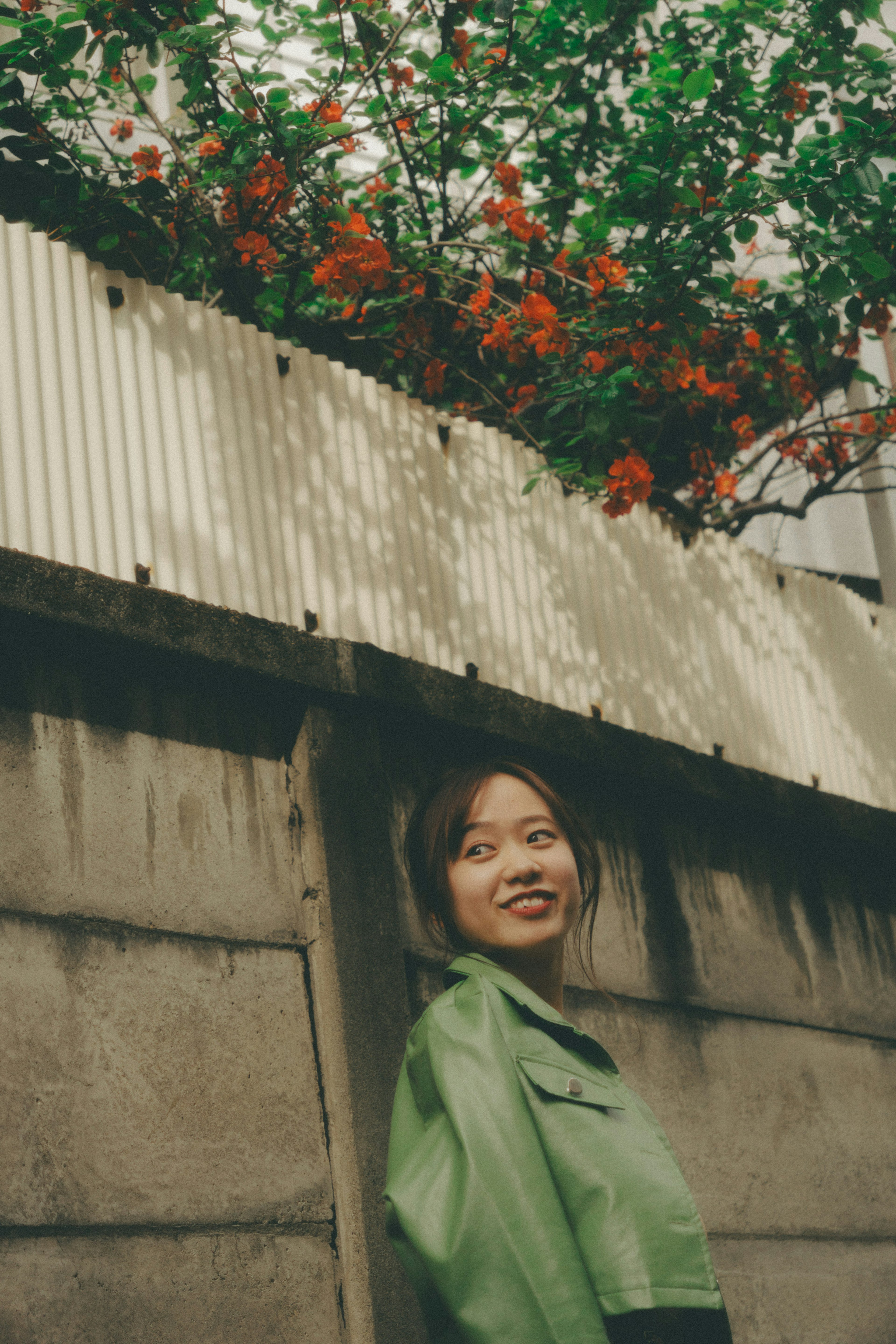 Woman in green jacket standing by a wall with orange flowering plants above