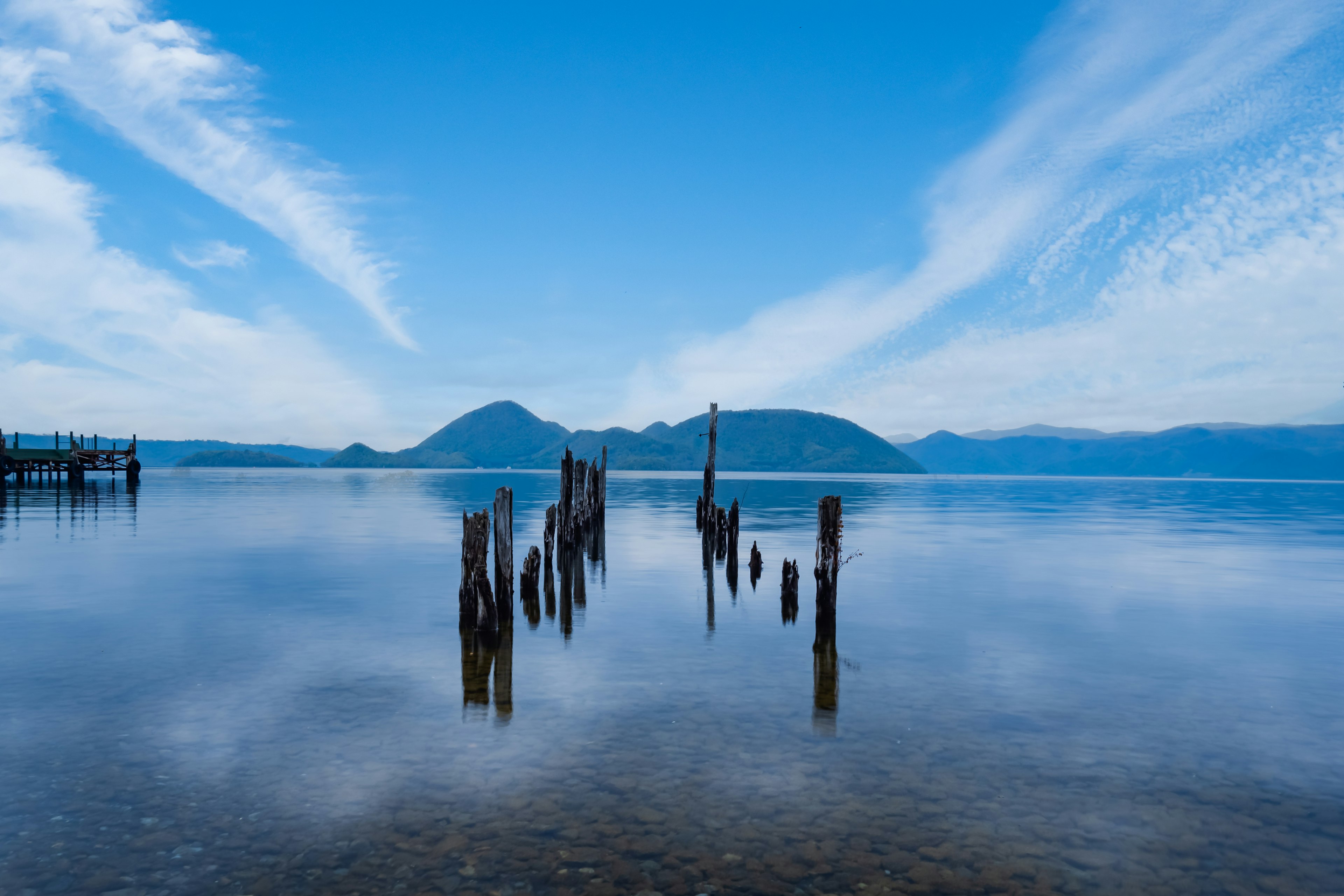 Old pier posts rising from calm lake water under a blue sky