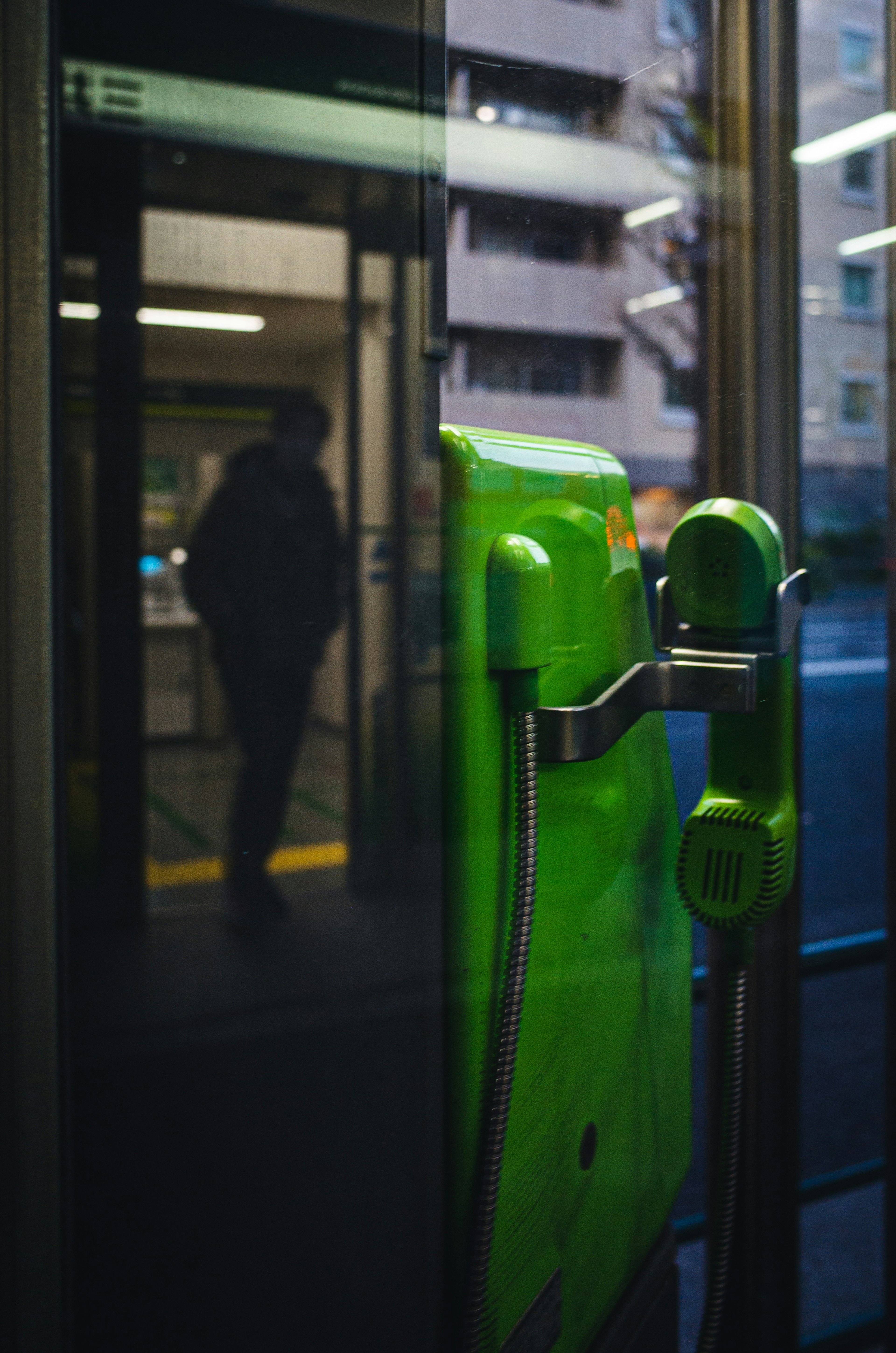 A green payphone reflected in glass with a silhouette of a person in the background