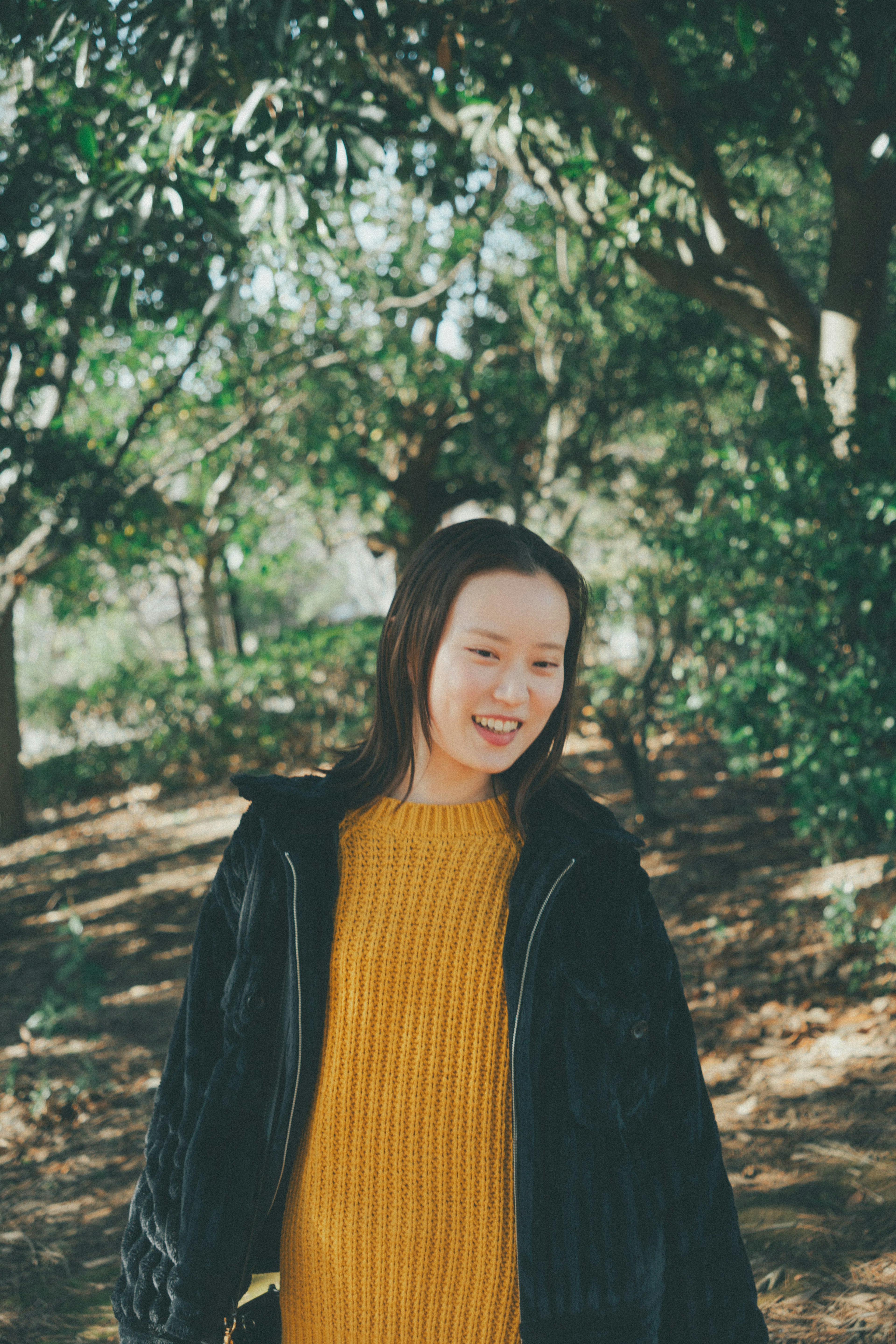 Femme souriante dans un parc portant un pull jaune et une veste noire entourée d'arbres verts