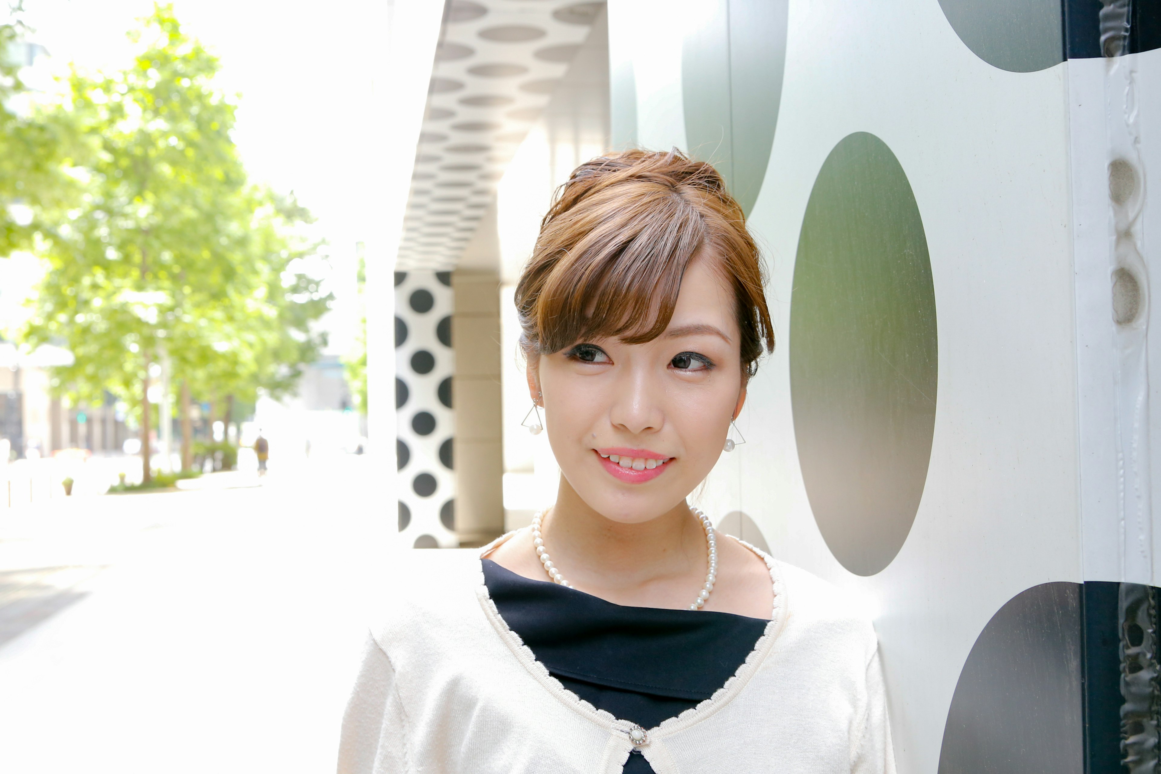 Portrait of a woman standing against a bright background wearing a black outfit with a white cardigan vibrant green trees and circular patterned wall