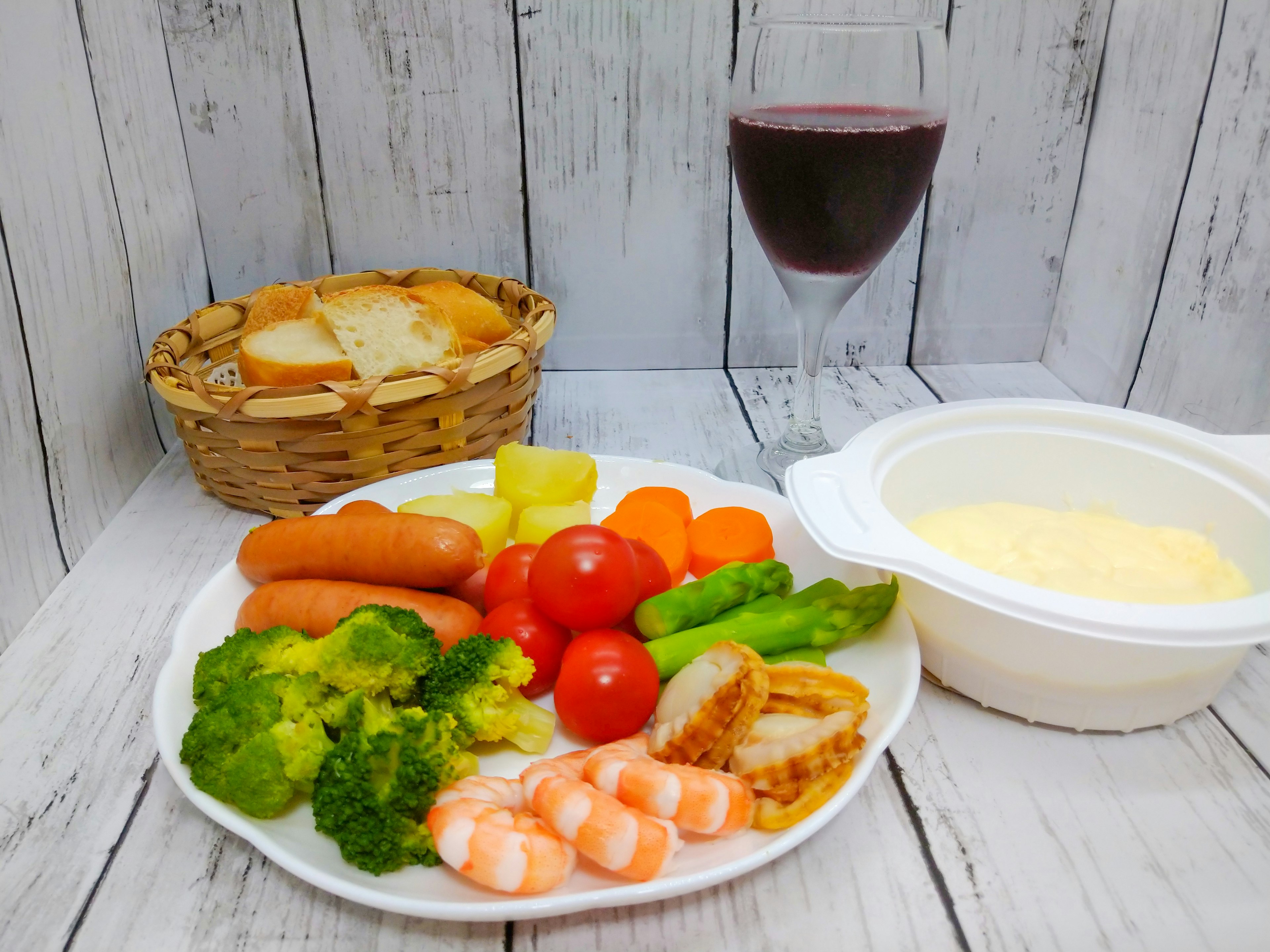 Plate with broccoli, tomatoes, shrimp, sausages, carrots, asparagus, grilled fish, and cream sauce alongside a glass of red wine and a basket of bread