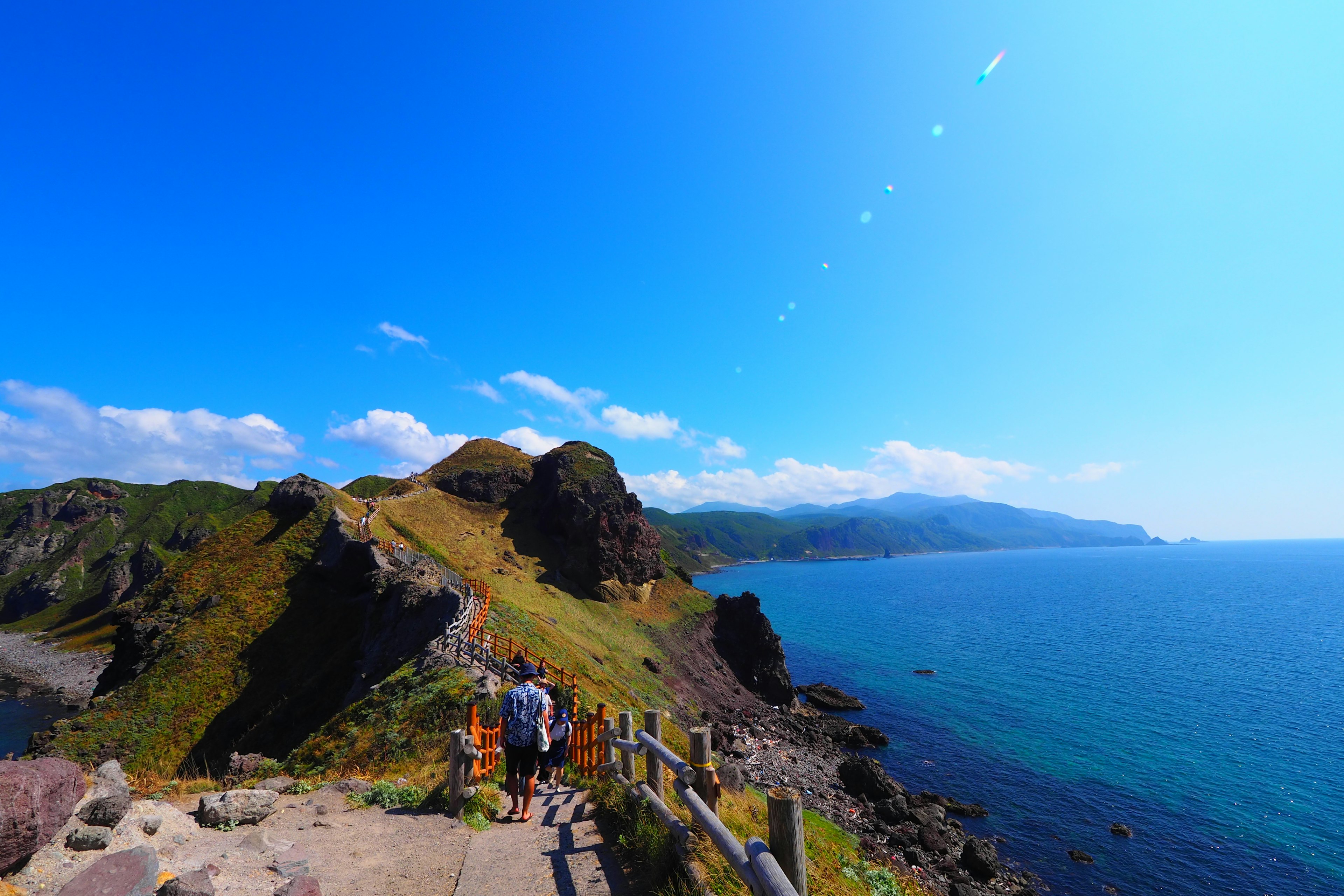 People walking along a mountain path with a backdrop of blue sea and sky