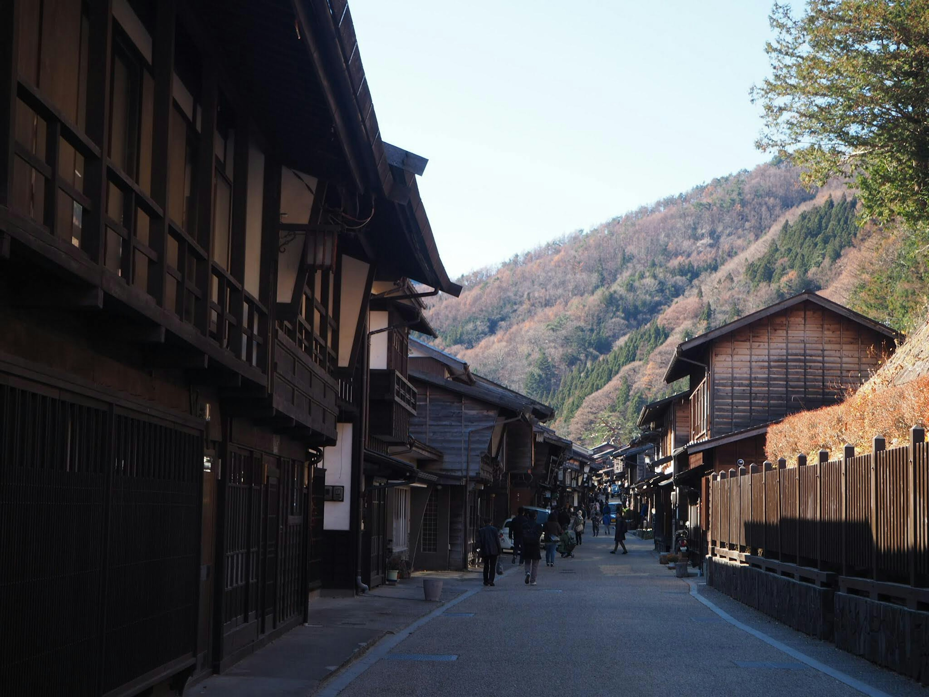 一幅寧靜的街景，展現了歷史悠久的日本城鎮中的傳統木建築，背景是山脈