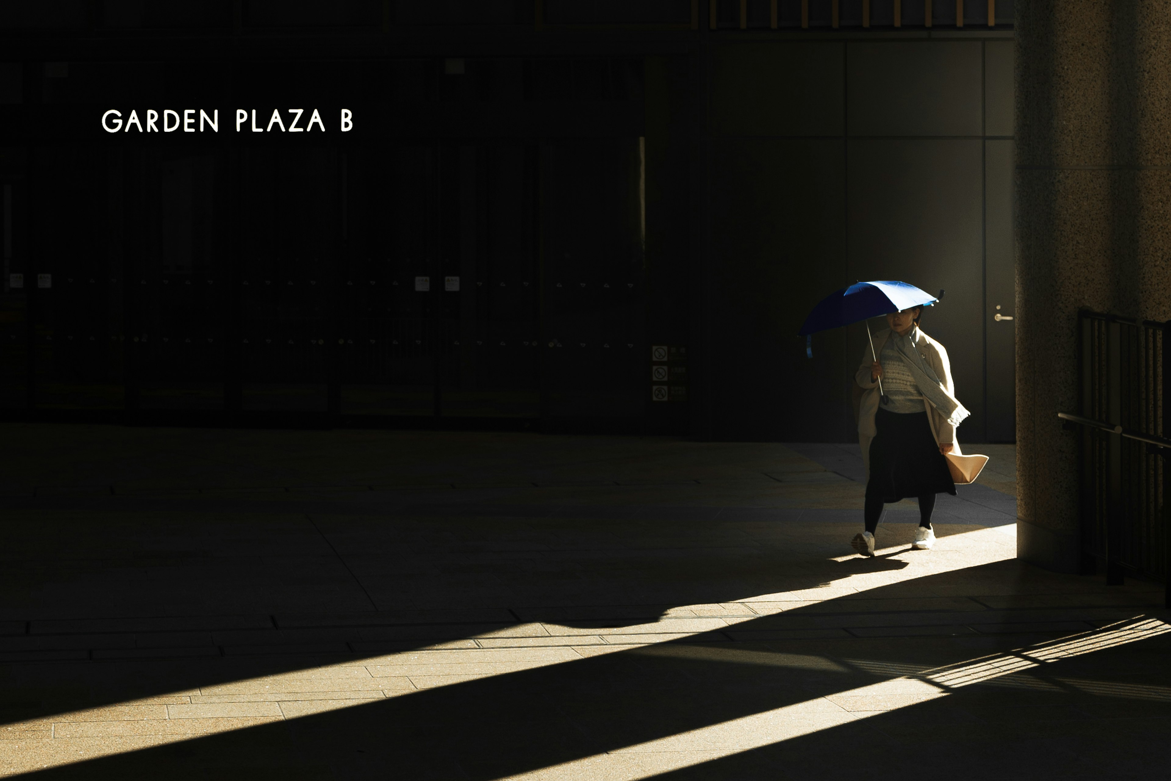 Woman walking with an umbrella in shadowy landscape sign for Garden Plaza B