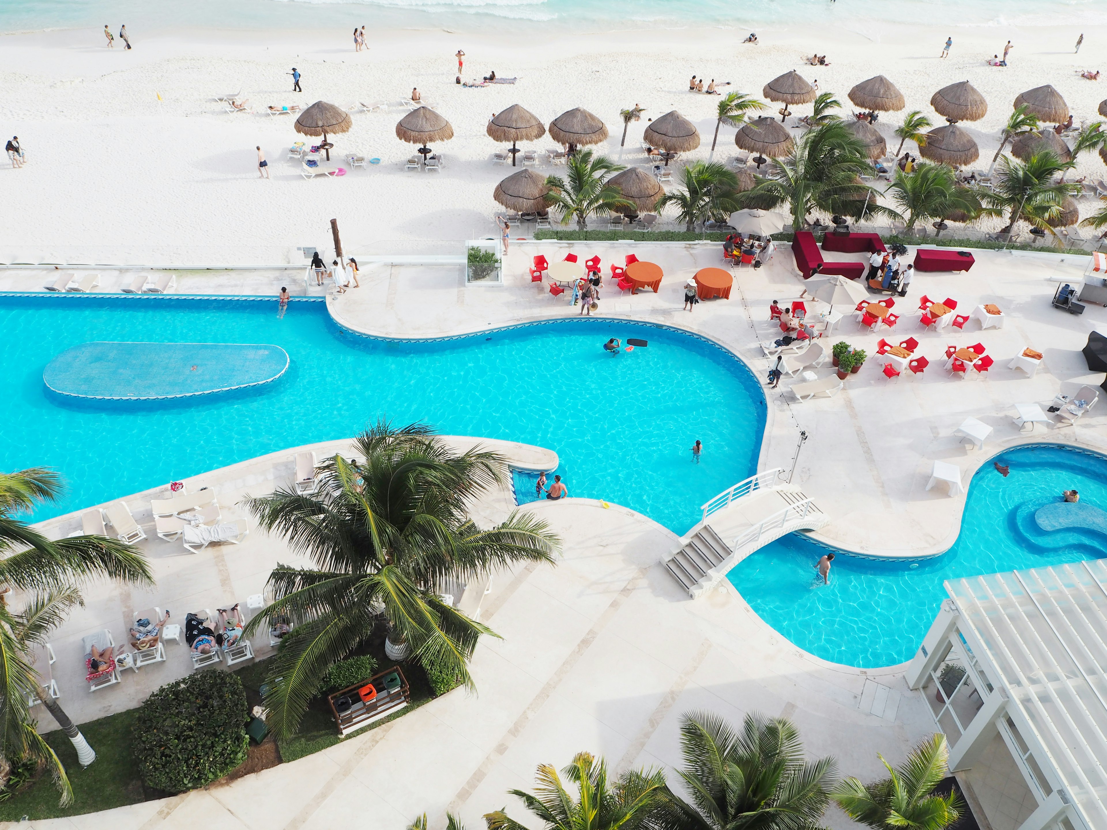 Scène de complexe avec piscine bleue et plage blanche avec des palmiers et parasols