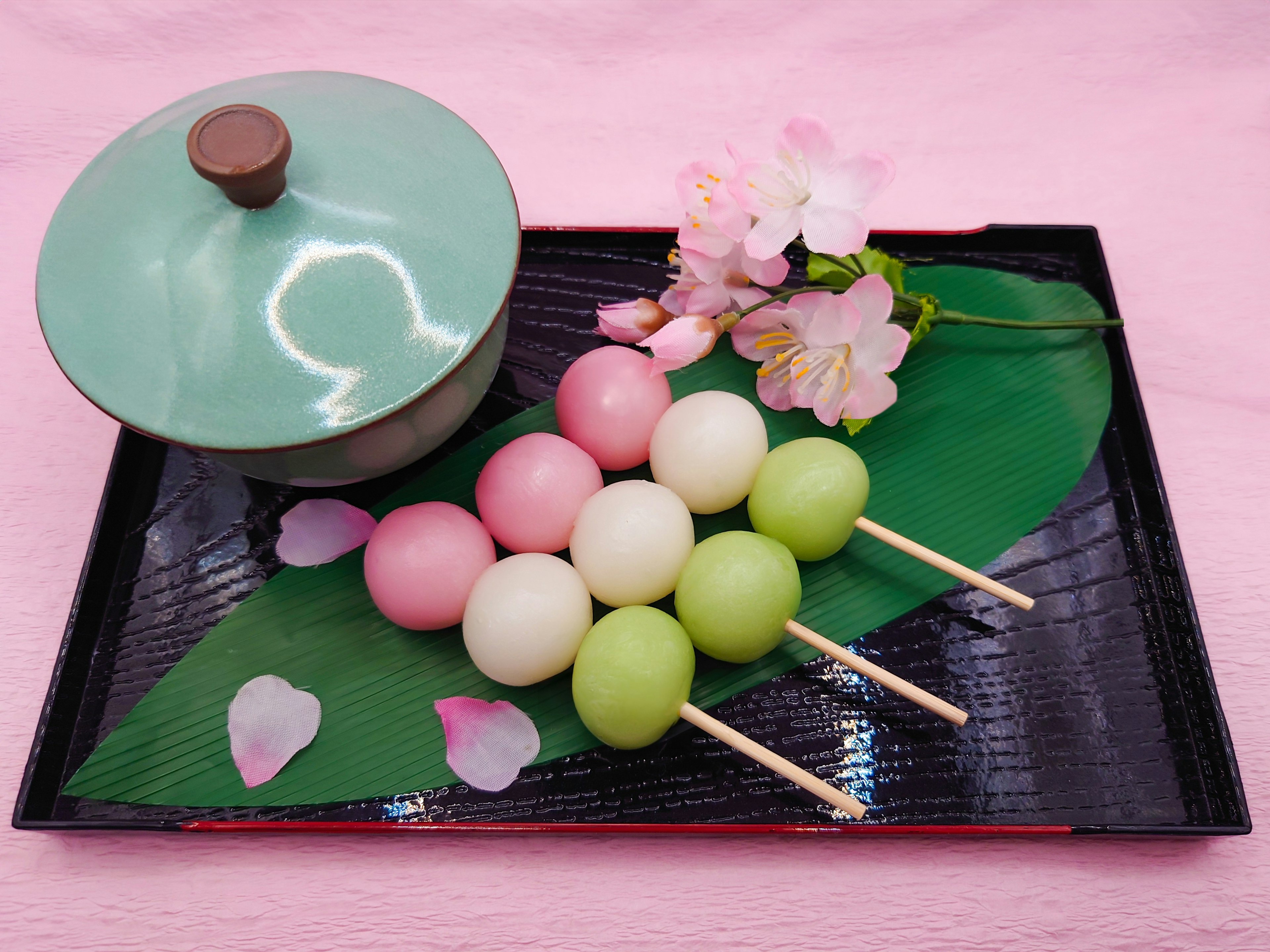 Brochettes de dango colorées disposées sur un plateau noir avec un bol vert et des fleurs de cerisier