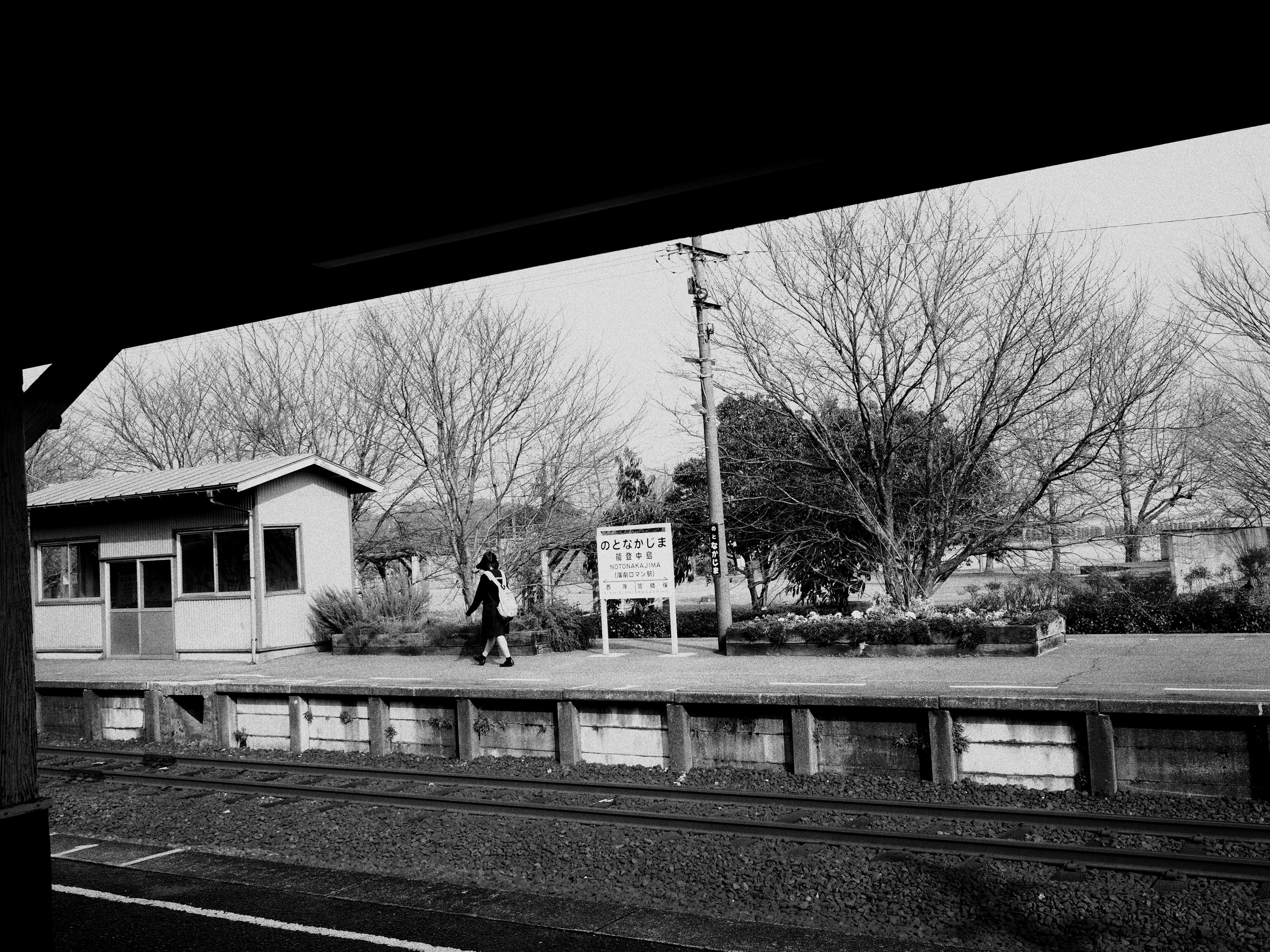 Schwarzweißfoto einer Person, die auf einem Bahnsteig mit Bäumen und einem Bahnhofgebäude geht