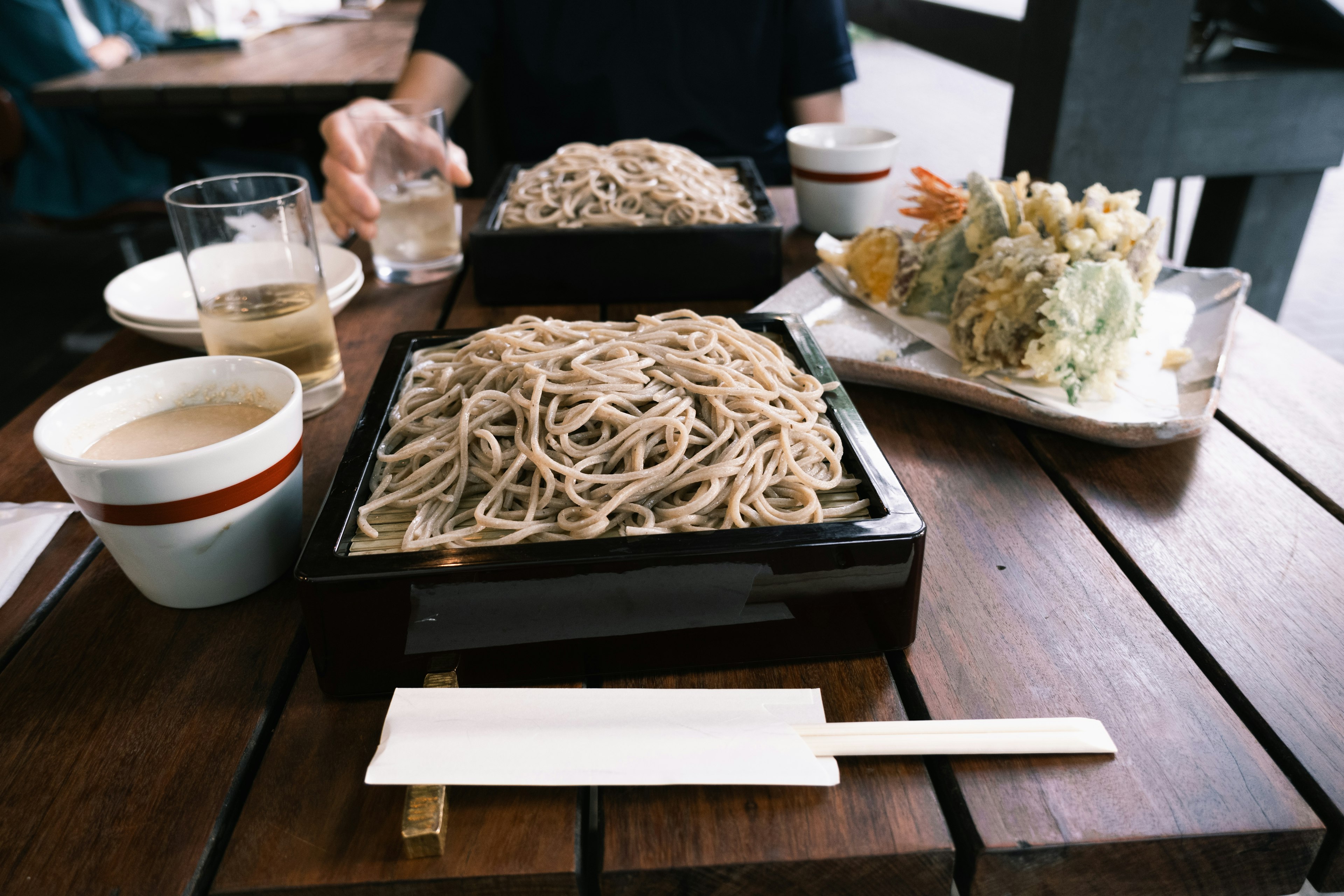 桌子上擺放著蕎麥麵和天婦羅菜餚，背景中有飲料和其他食物