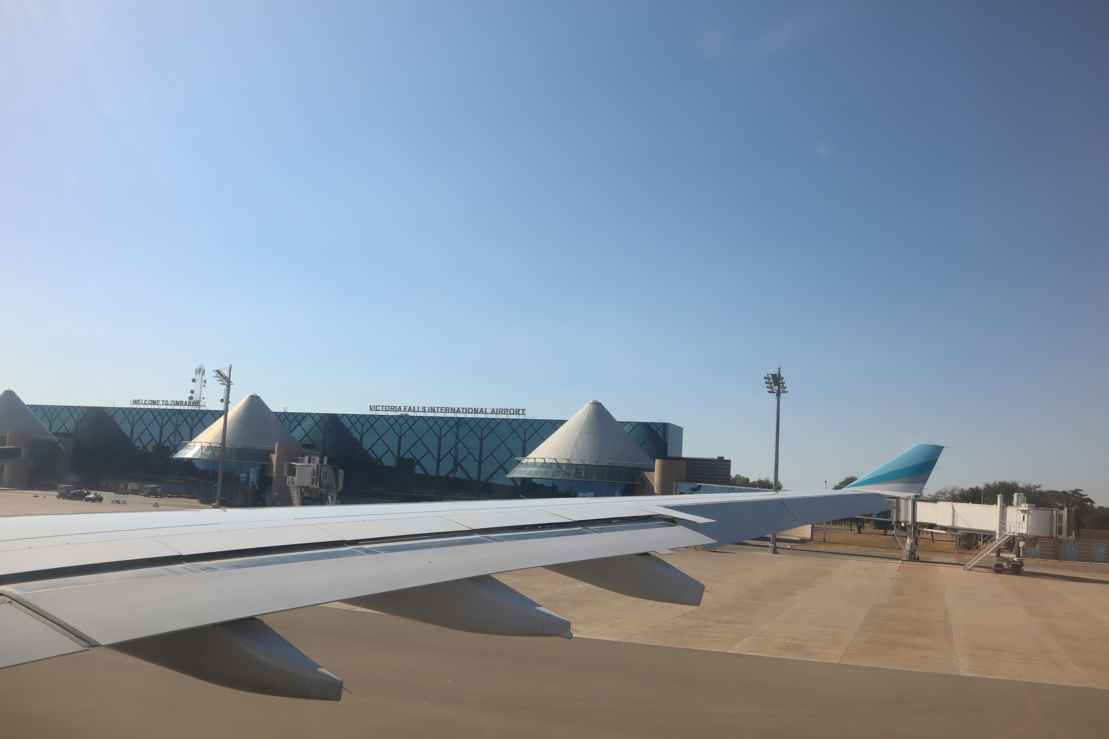 Pemandangan sayap pesawat dengan bangunan bandara di latar belakang dan langit biru yang cerah