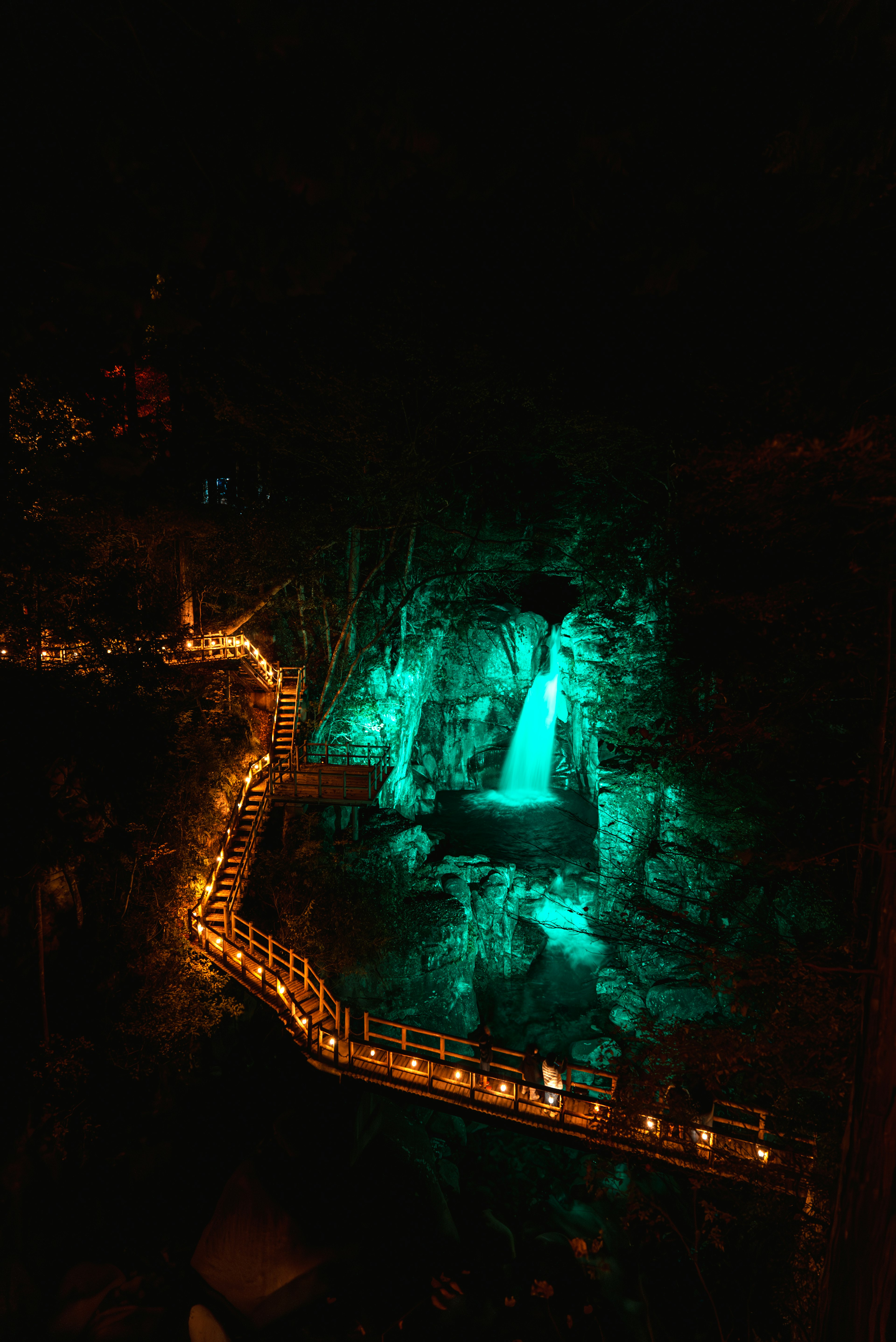 Nächtlicher Blick auf einen teal beleuchteten Wasserfall mit einem gewundenen Weg