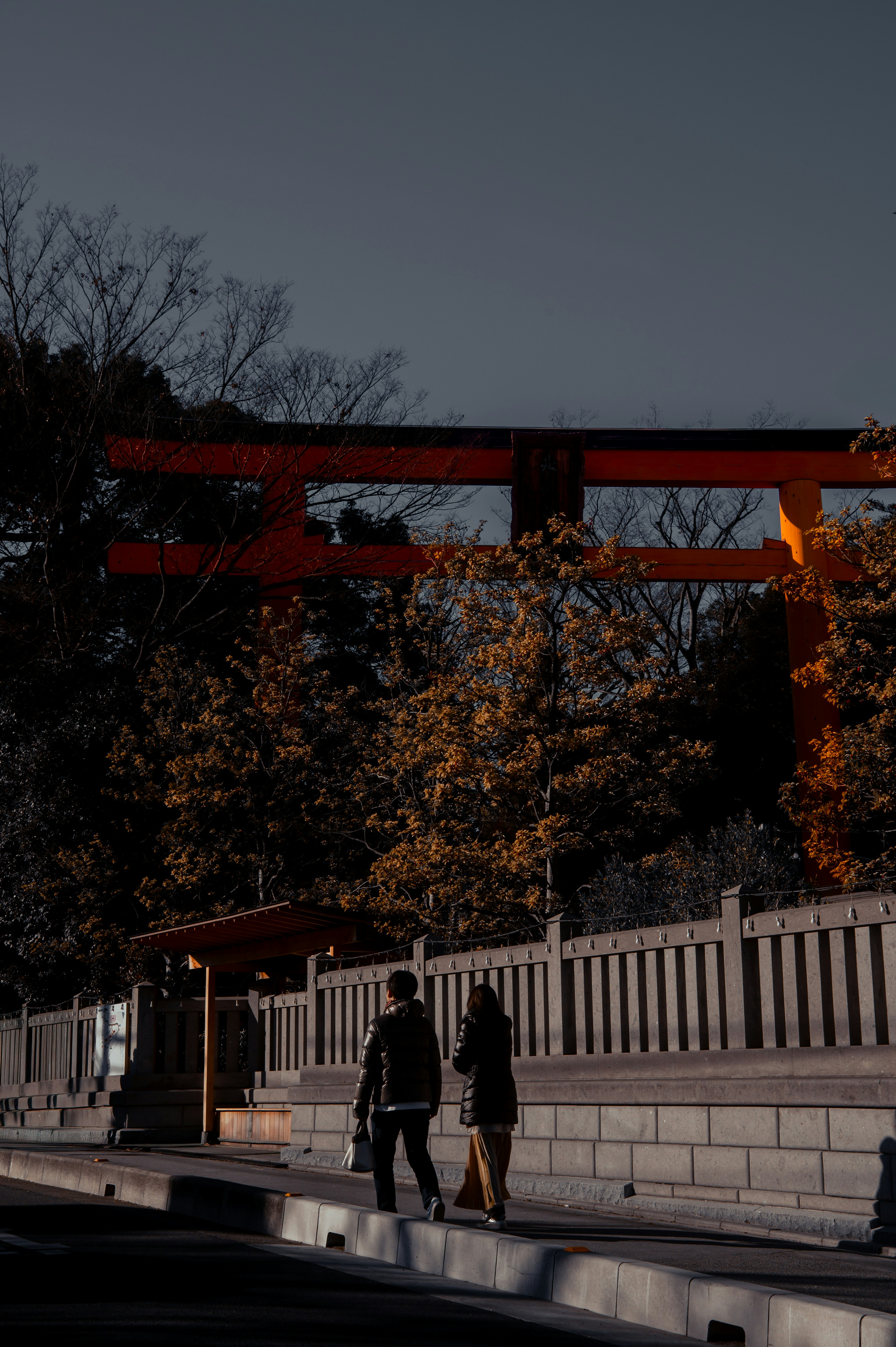 Silueta de dos personas caminando bajo un torii rojo al anochecer