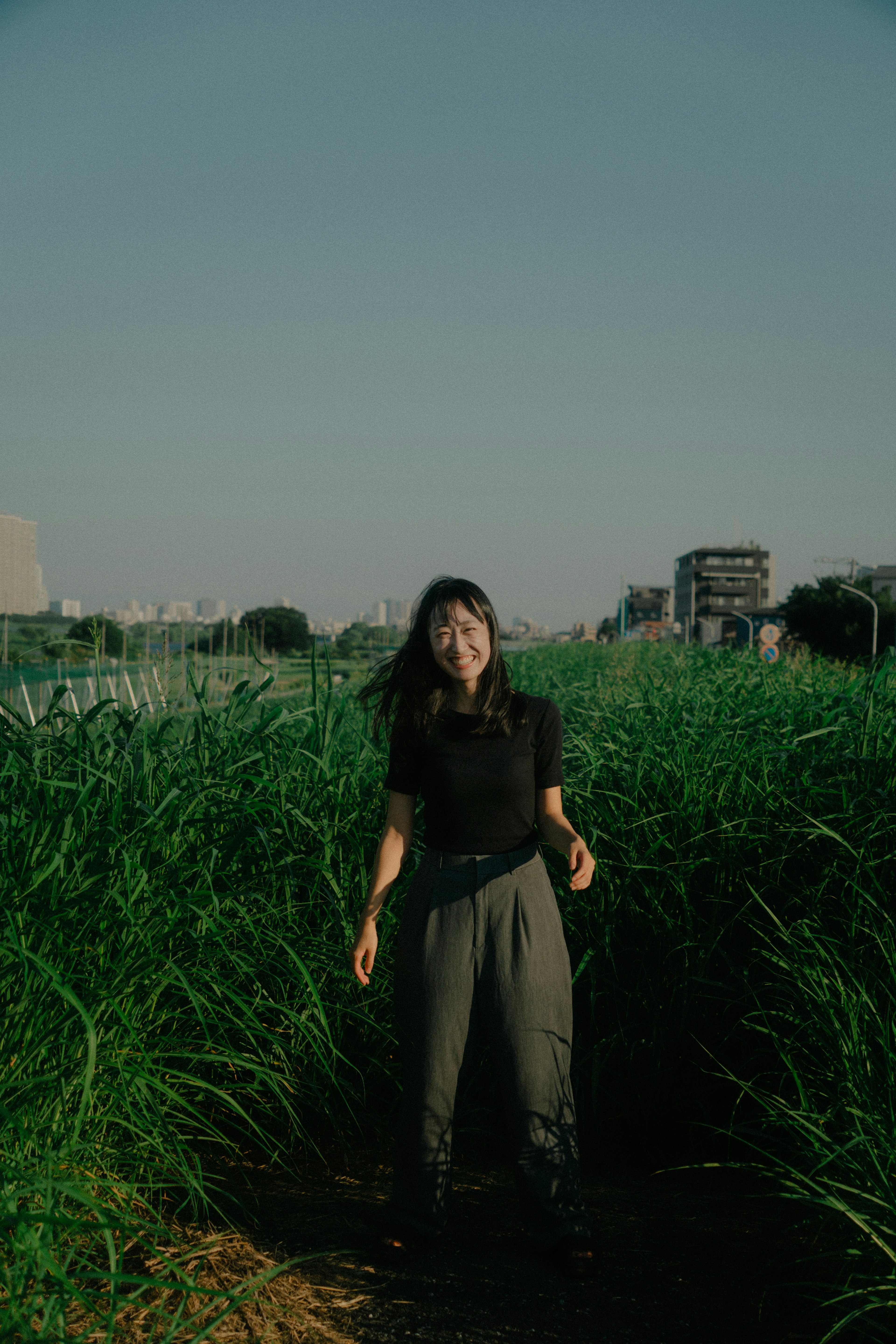 A woman posing amidst tall green grass with buildings in the background