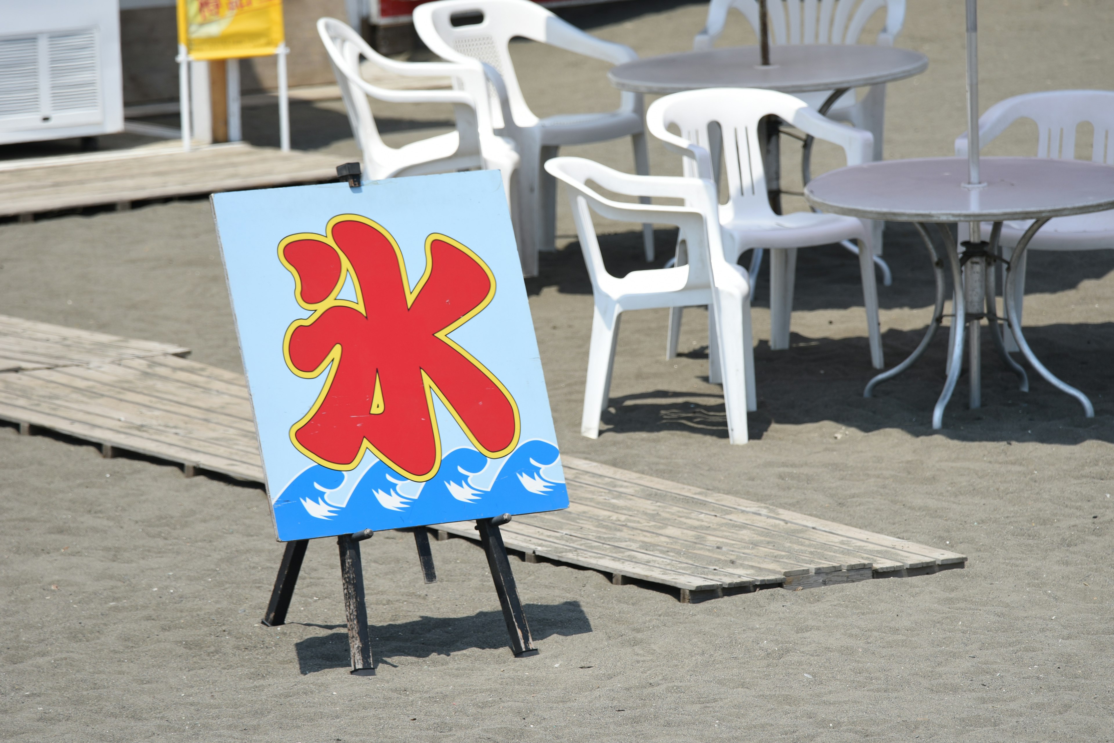 A blue sign with red ice characters near white chairs and tables on the beach