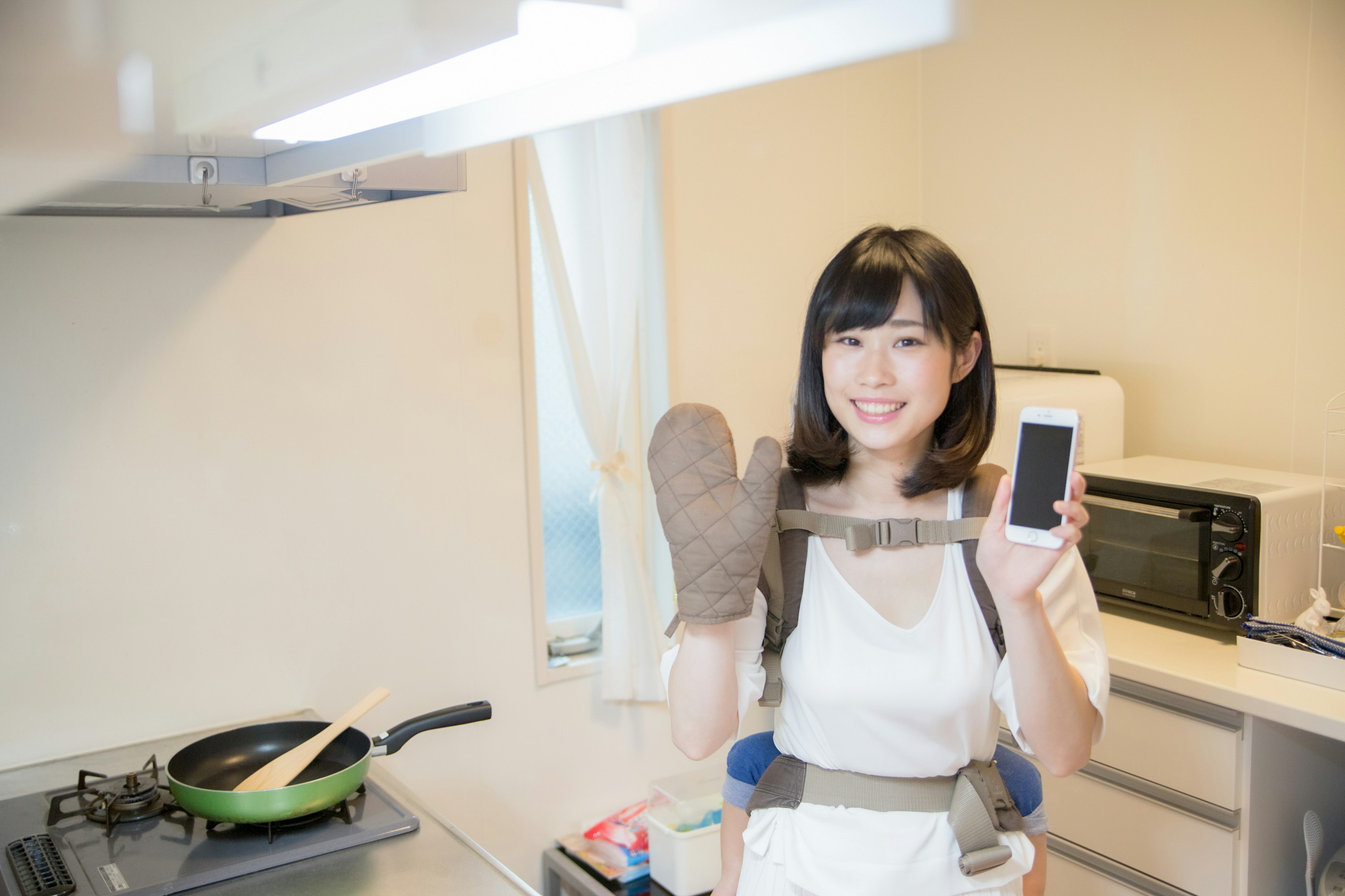 Smiling woman in kitchen wearing oven mitts holding smartphone