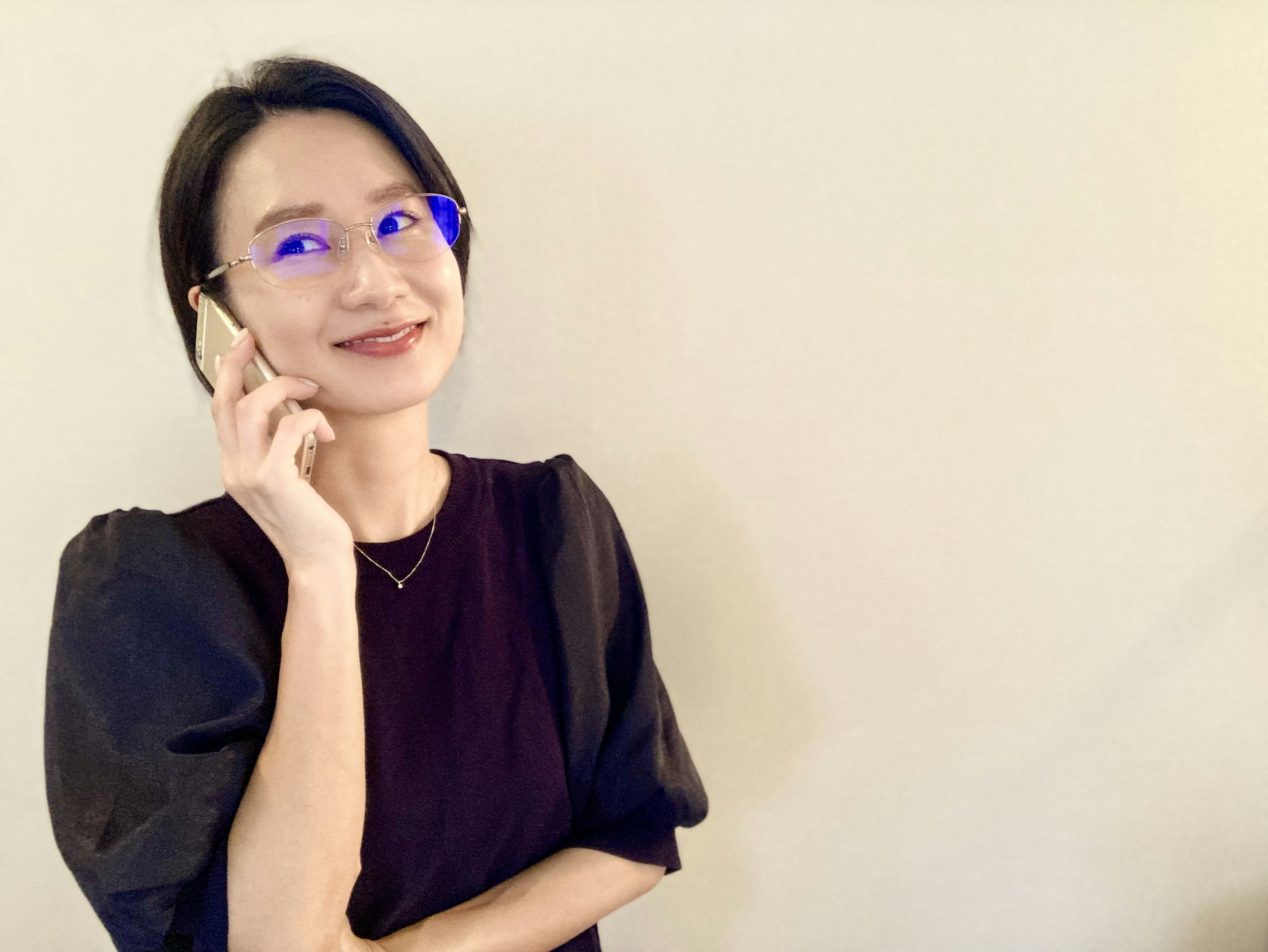 Portrait d'une femme souriante au téléphone portant une tenue noire et des lunettes bleues avec un fond crème simple