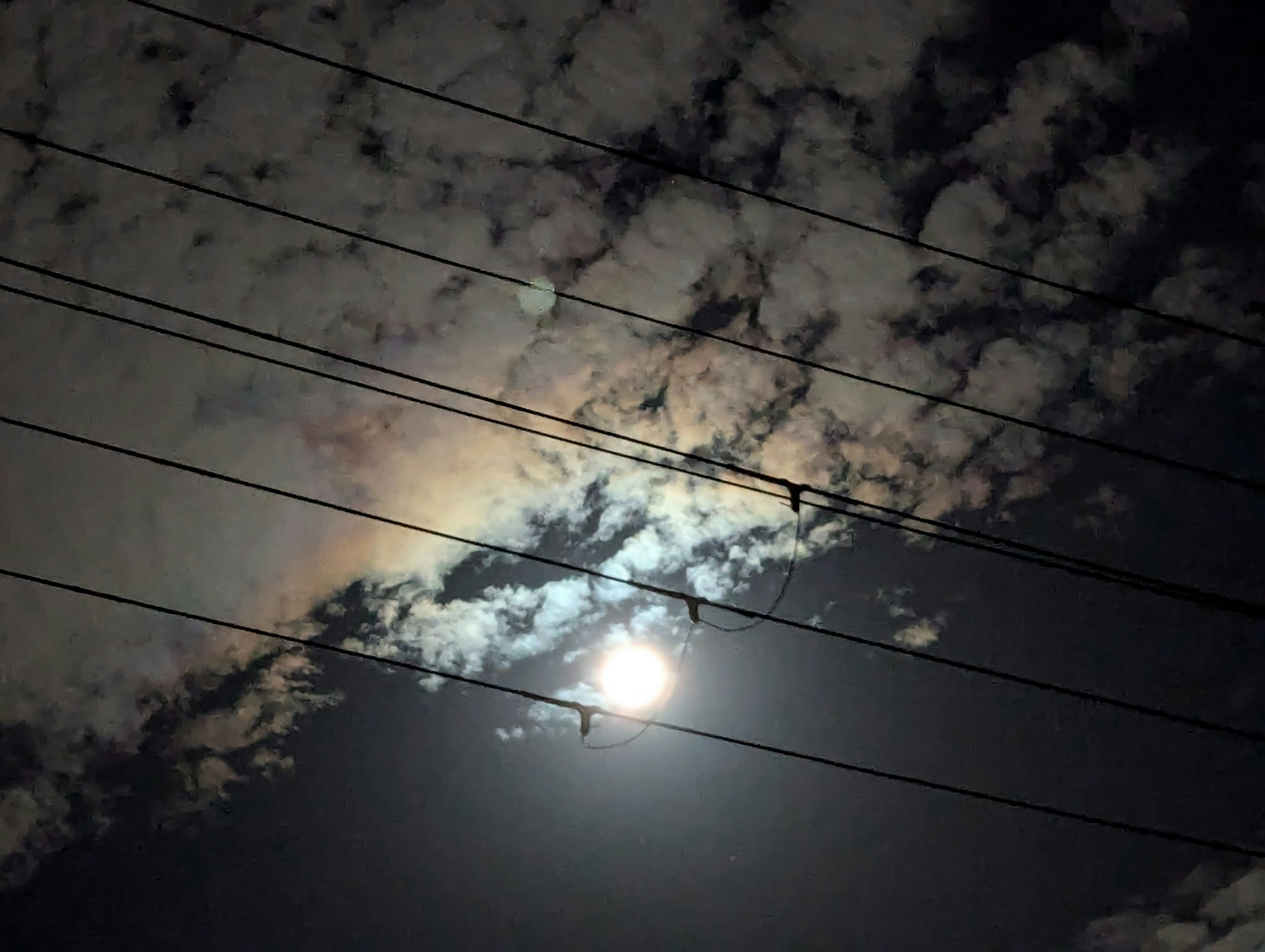 A dark night sky featuring the moon and clouds with power lines in the foreground