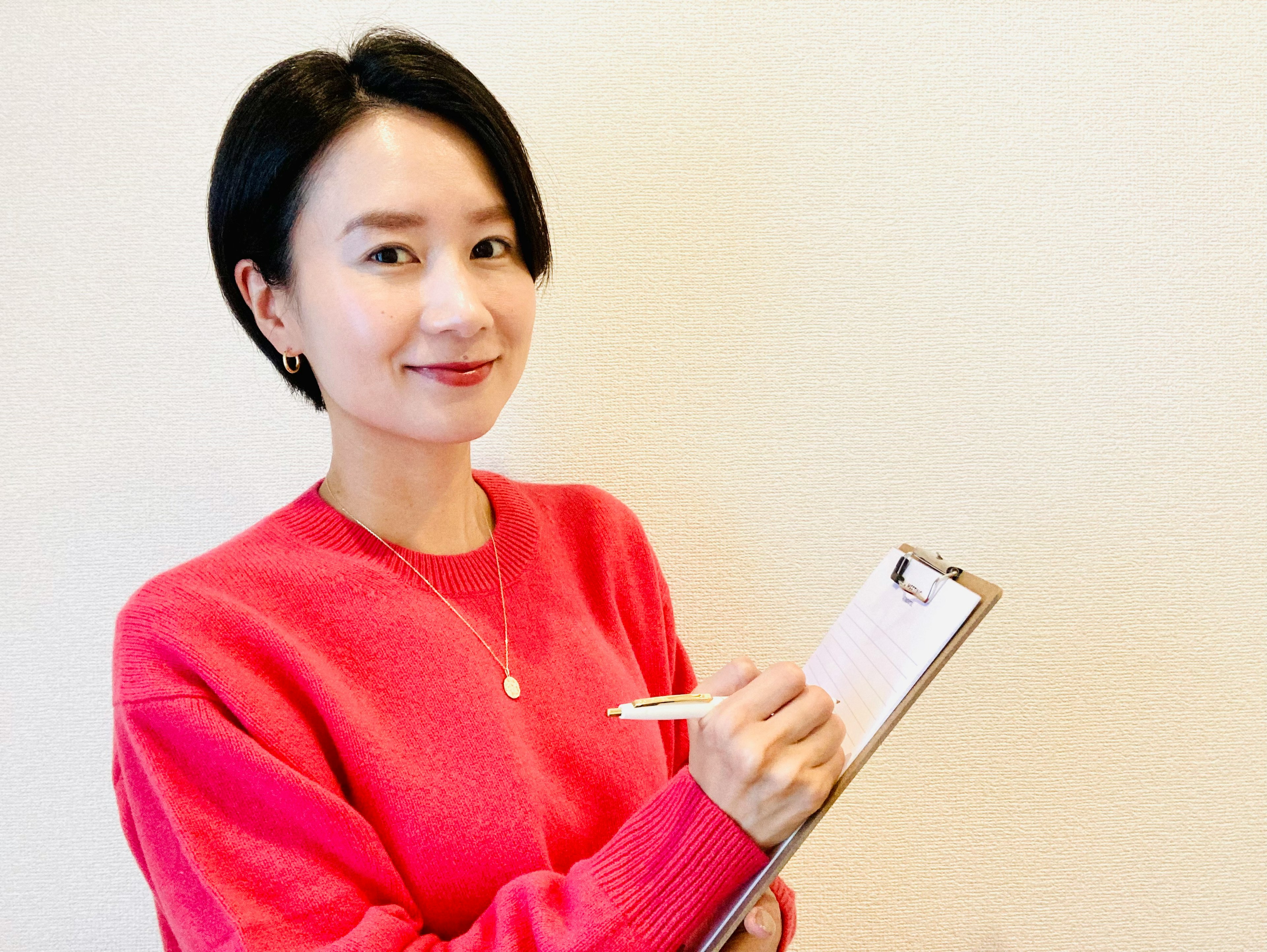 A woman wearing a pink sweater holding a clipboard and pen smiling