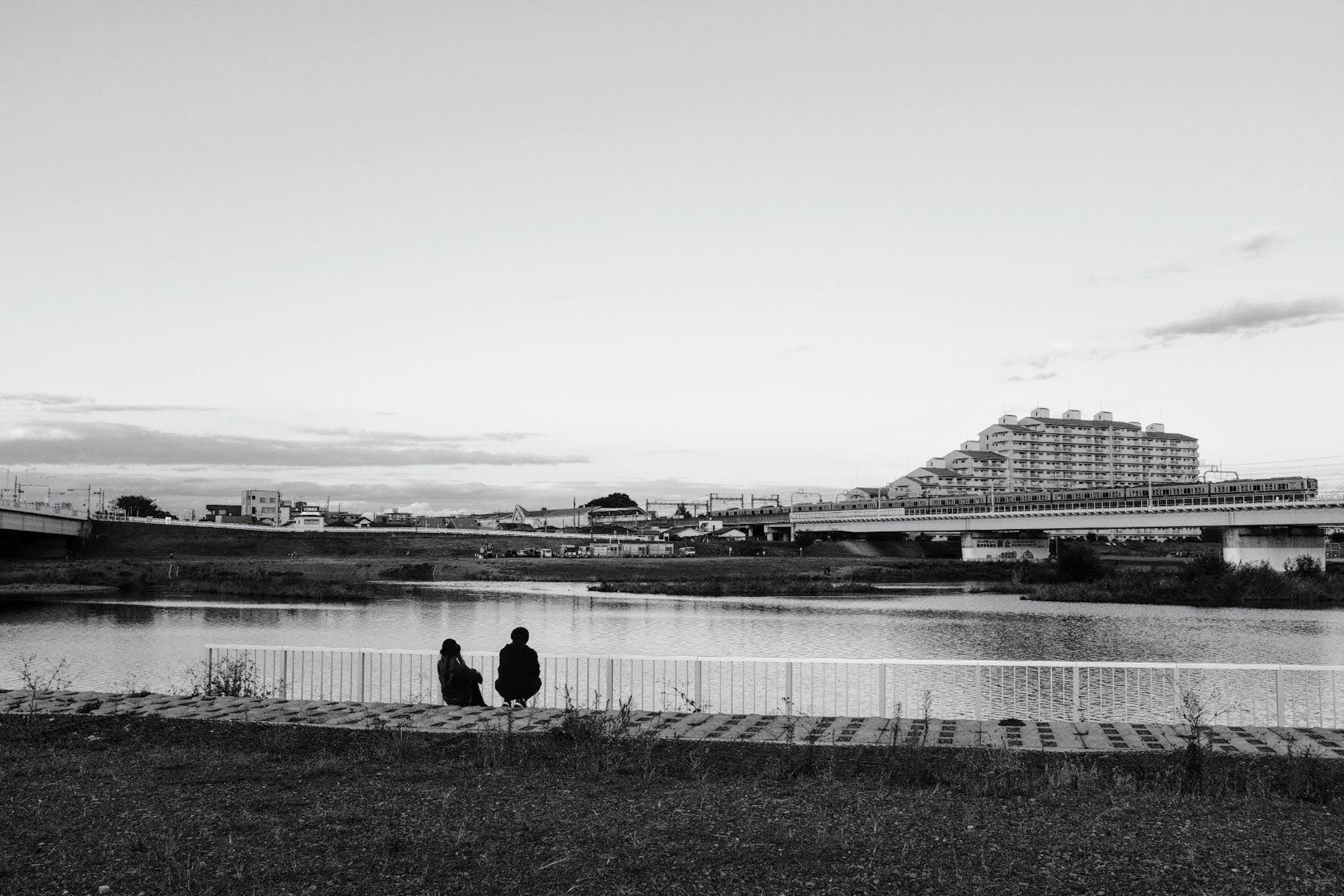 兩個人坐在河邊，背後是建築物