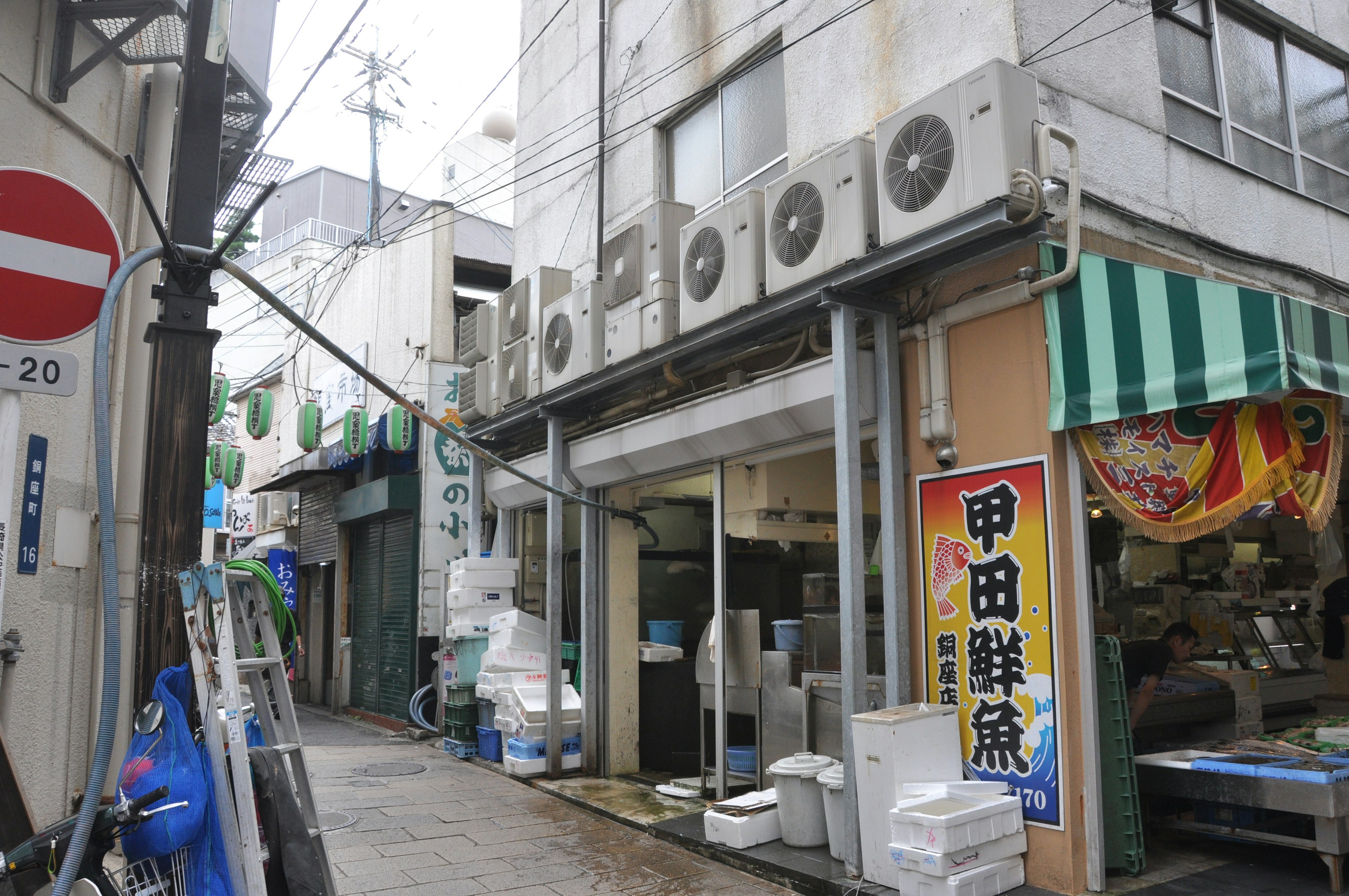 A view of a narrow street with shops featuring air conditioning units on the exterior and a sign in Japanese indicating fresh fish