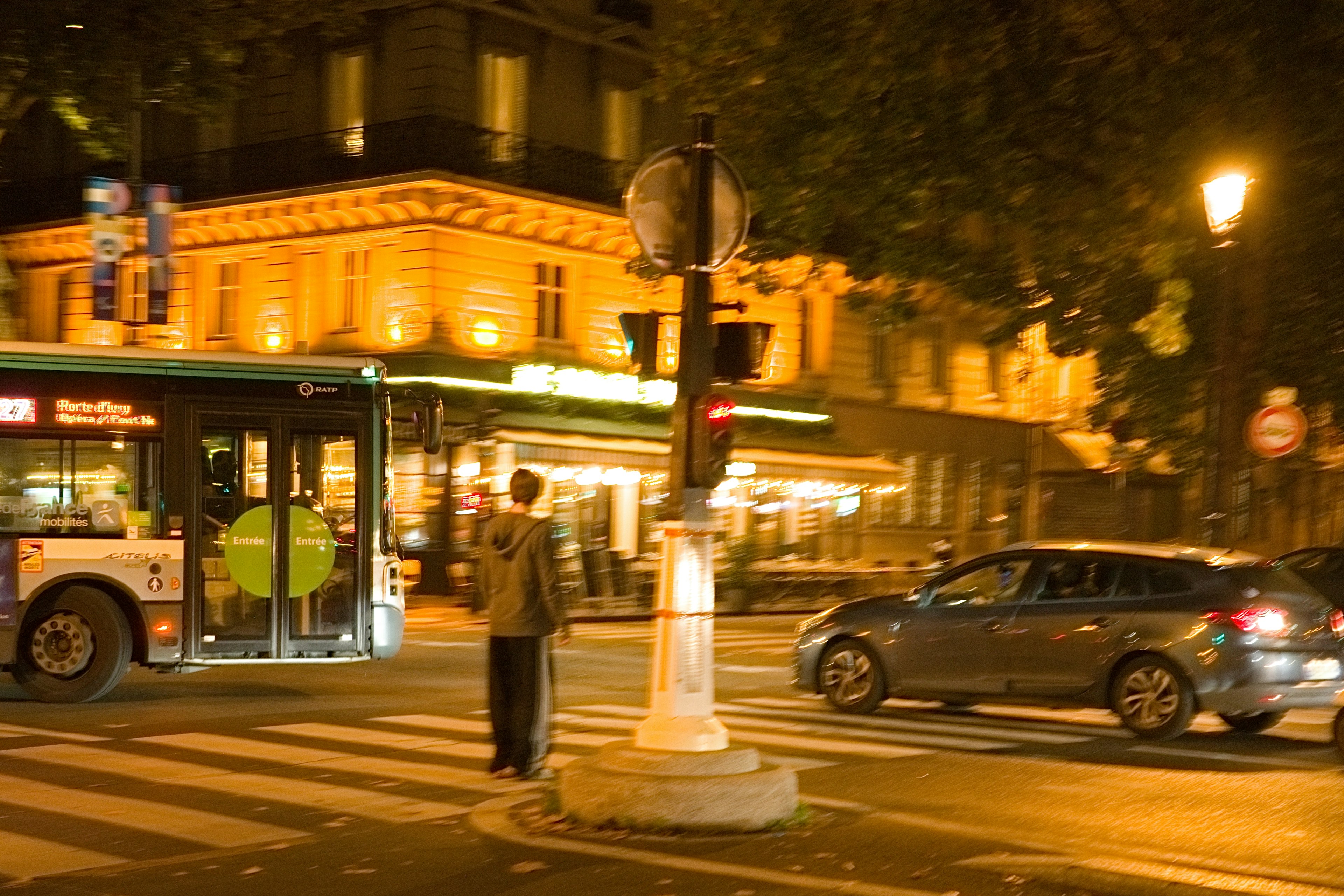 A person waiting at a traffic signal near a bus at night