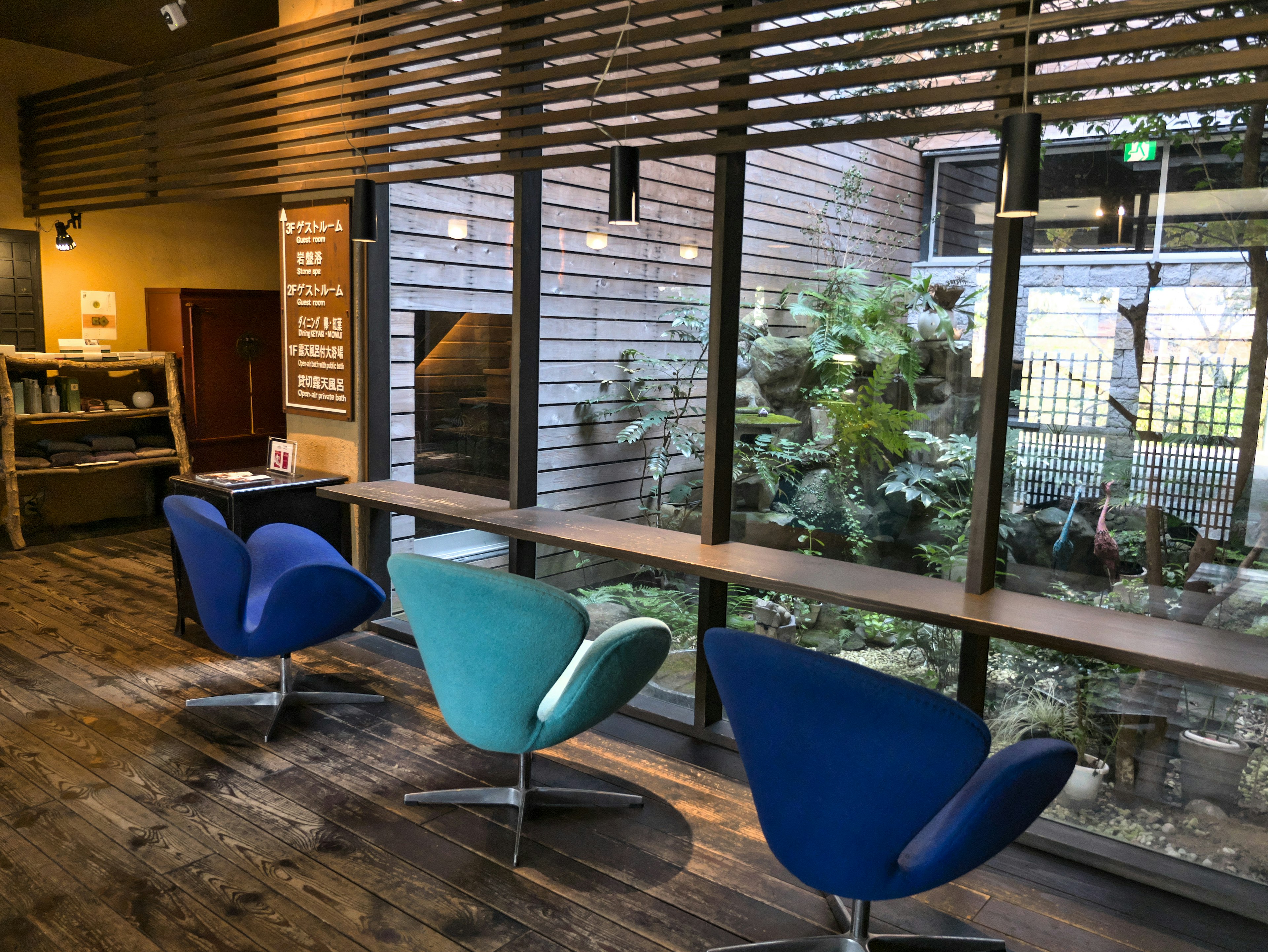 Interior de un café con suelo de madera grandes ventanas tres sillas coloridas y vegetación visible