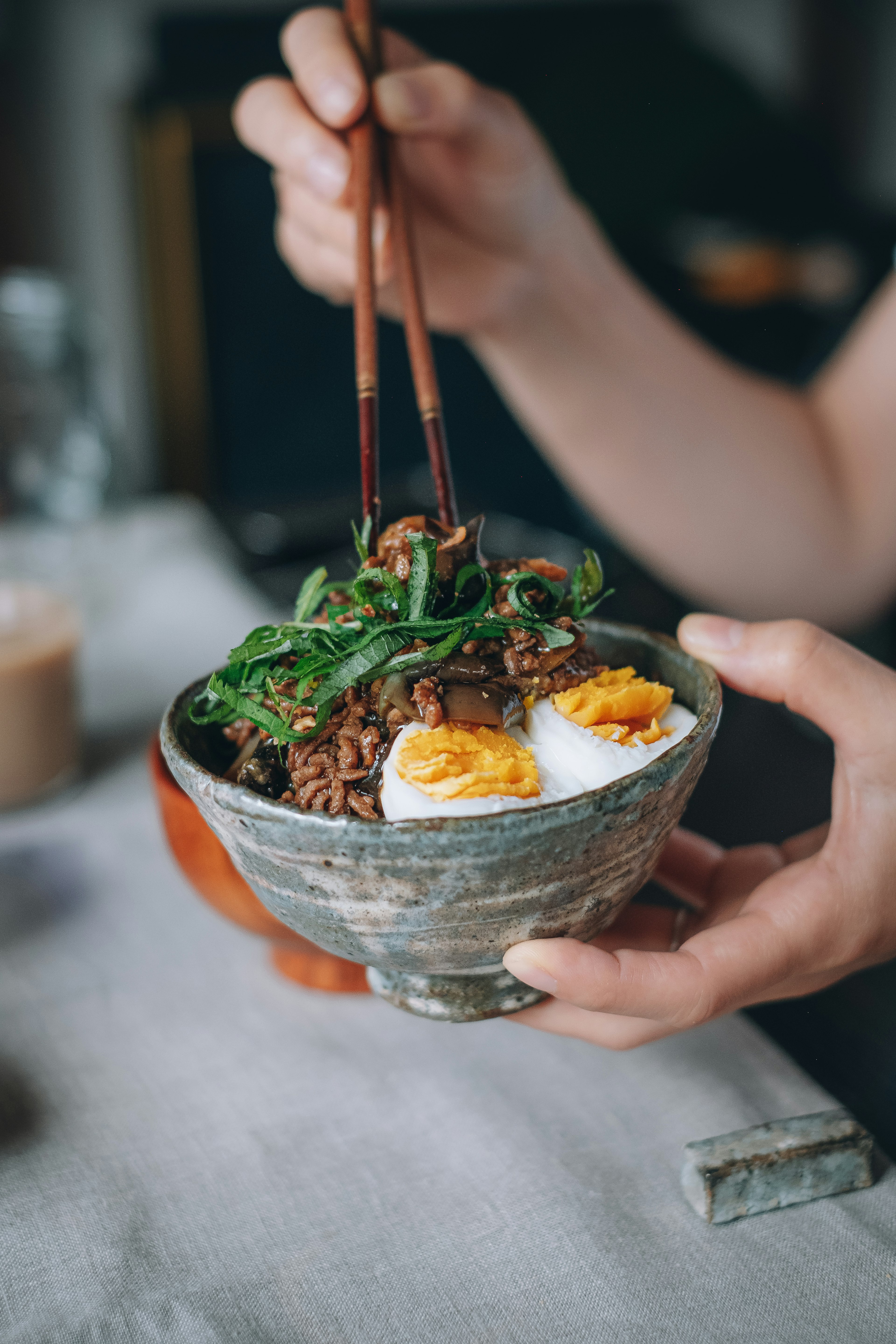 A hand holding a brown bowl filled with rice, meat, and soft-boiled eggs