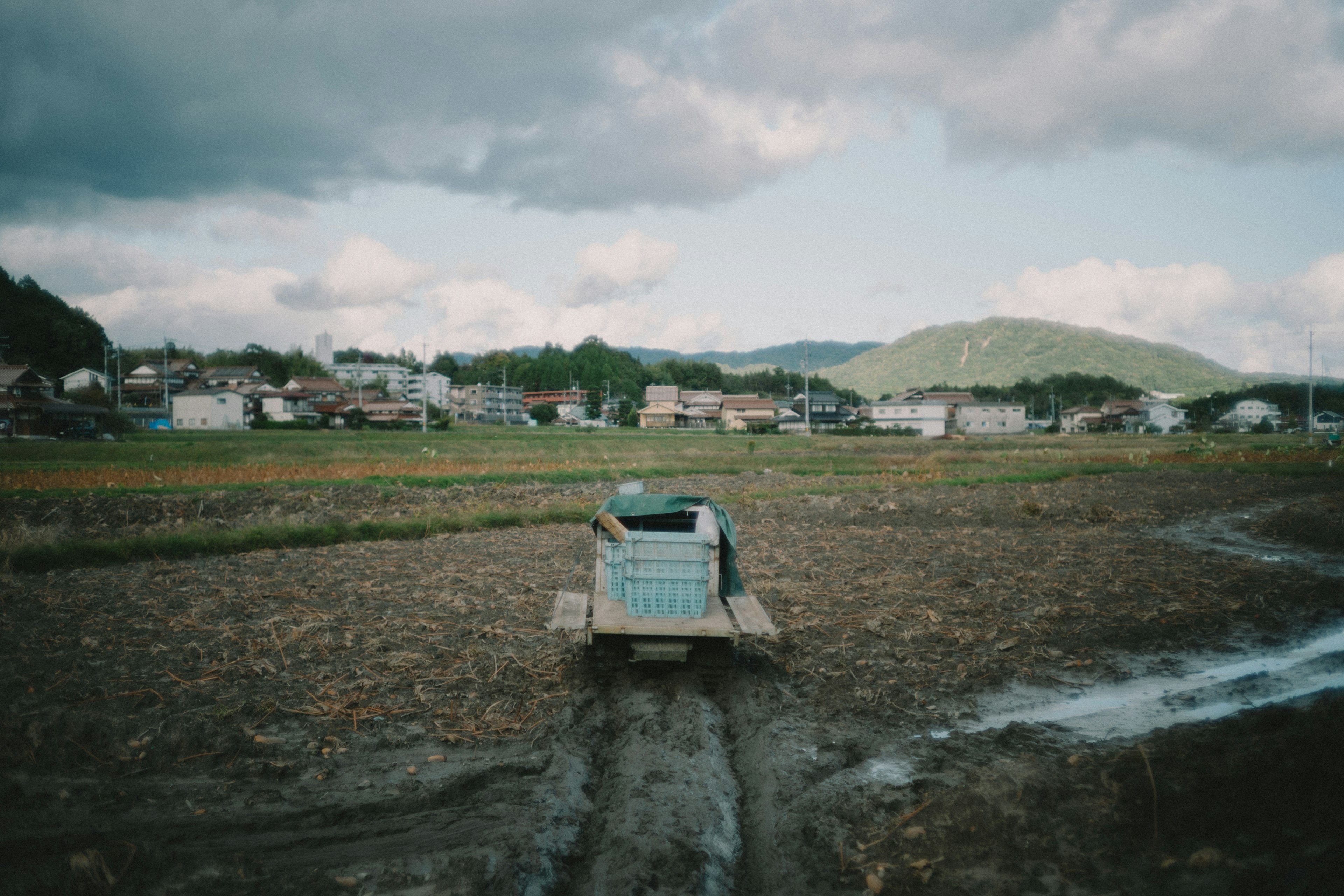 Traktor biru di tengah sawah dengan desa dan pegunungan di latar belakang