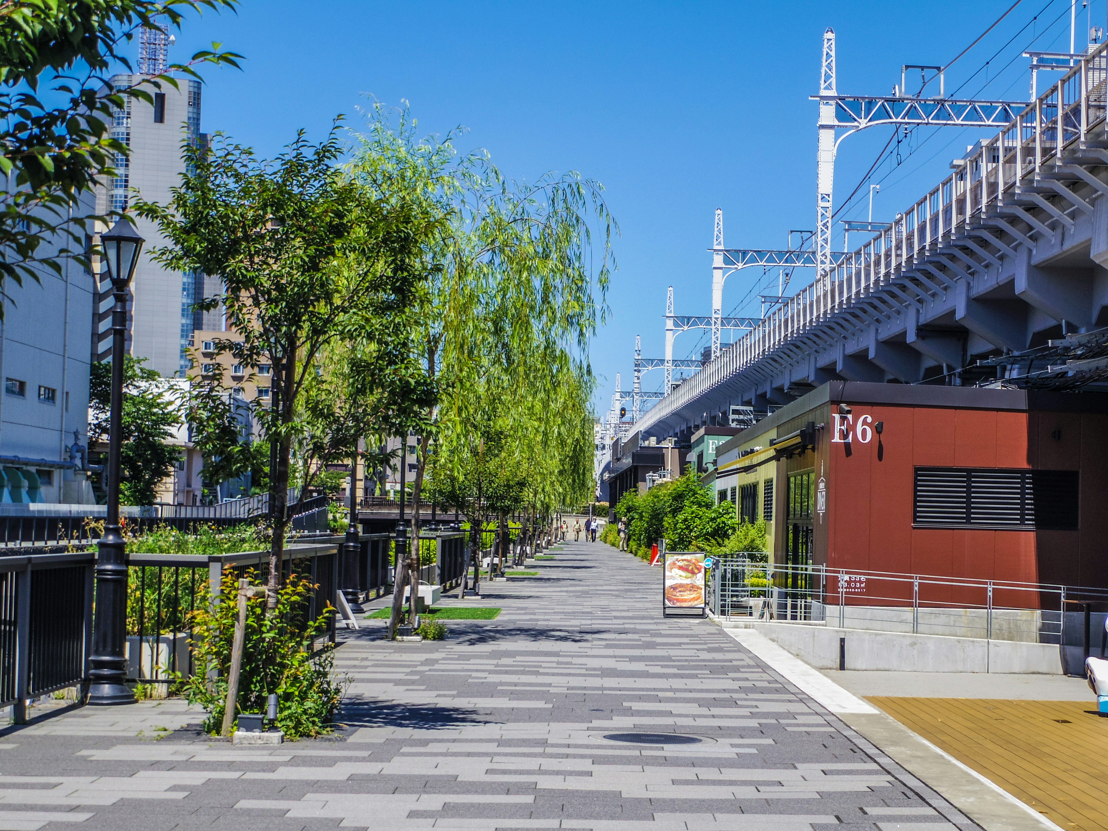 A vibrant walkway lined with trees under a bright blue sky featuring buildings and railway tracks