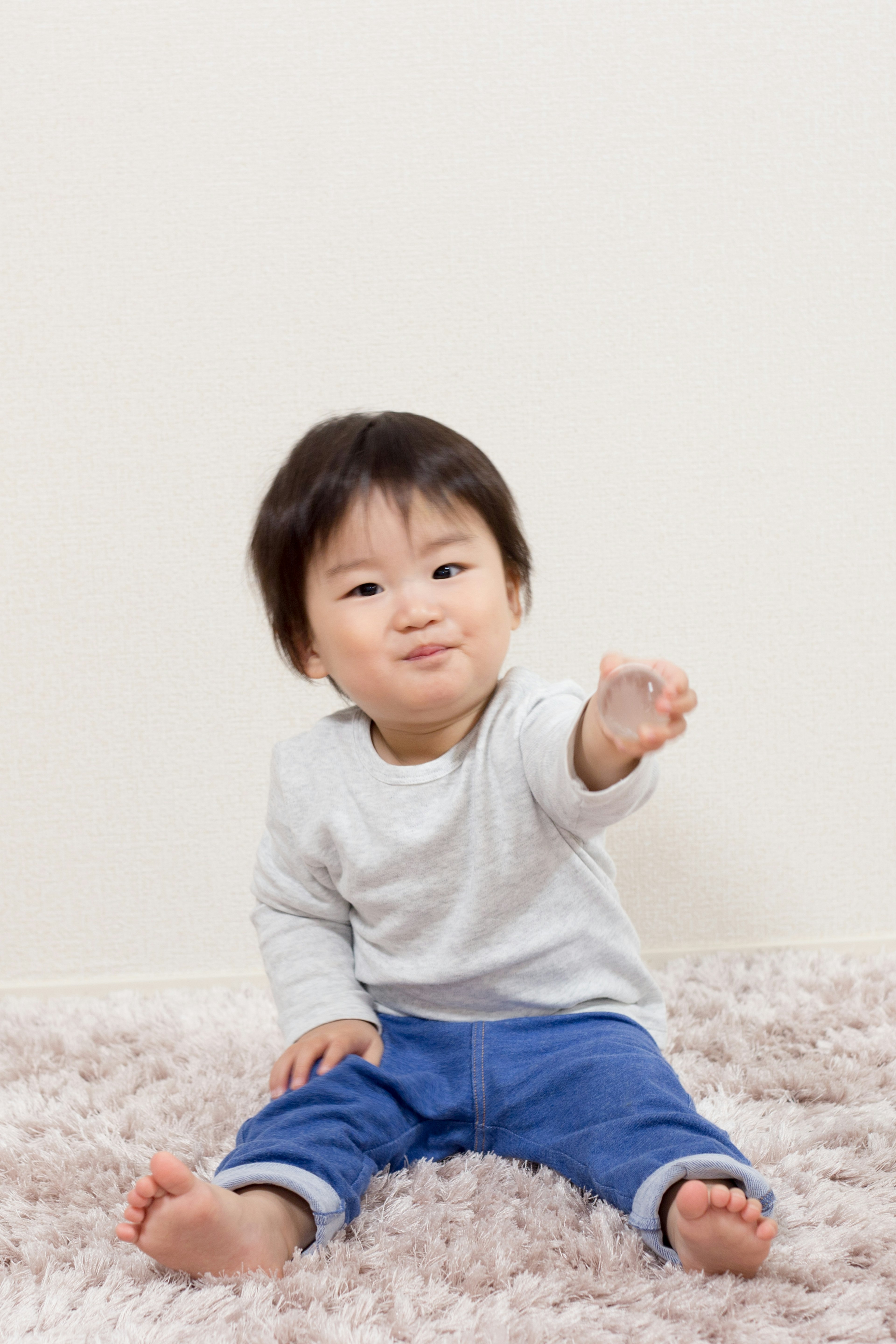 A small boy sitting on a soft rug in front of a white wall reaching out his hand