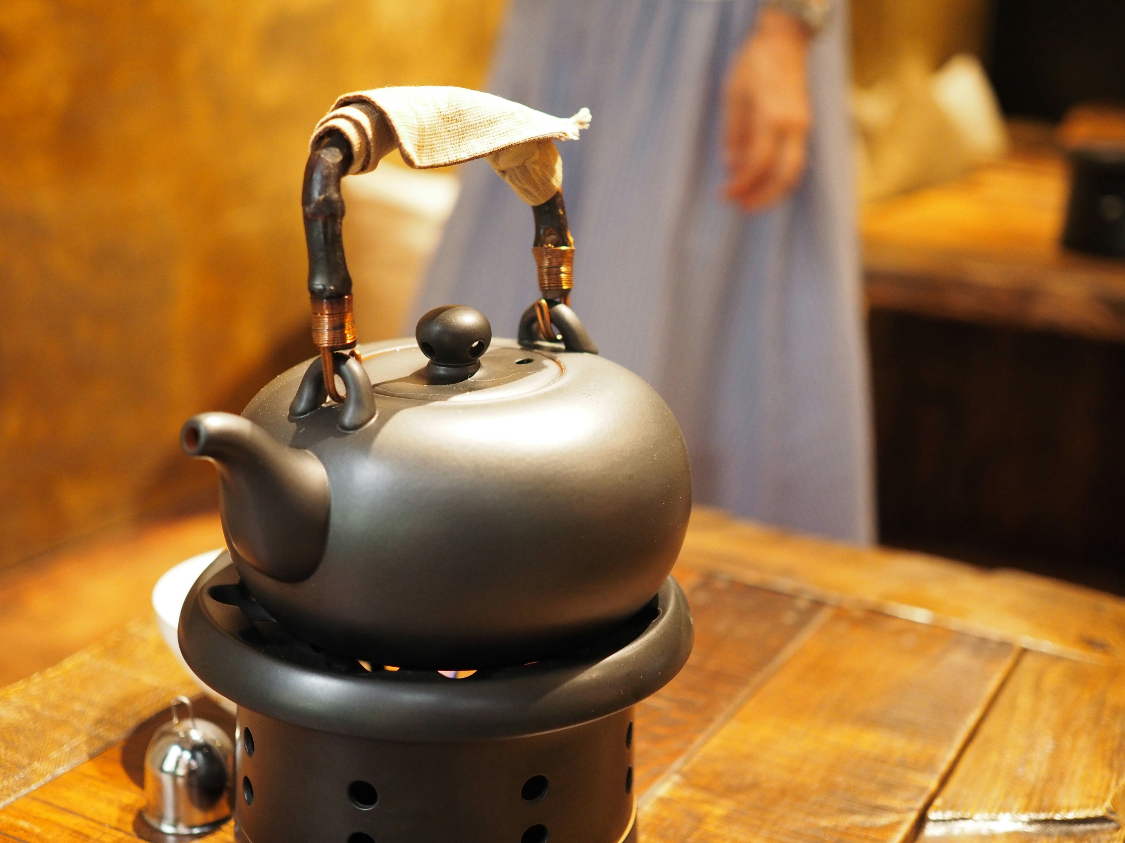 A black teapot on a wooden table with a blurred person in the background