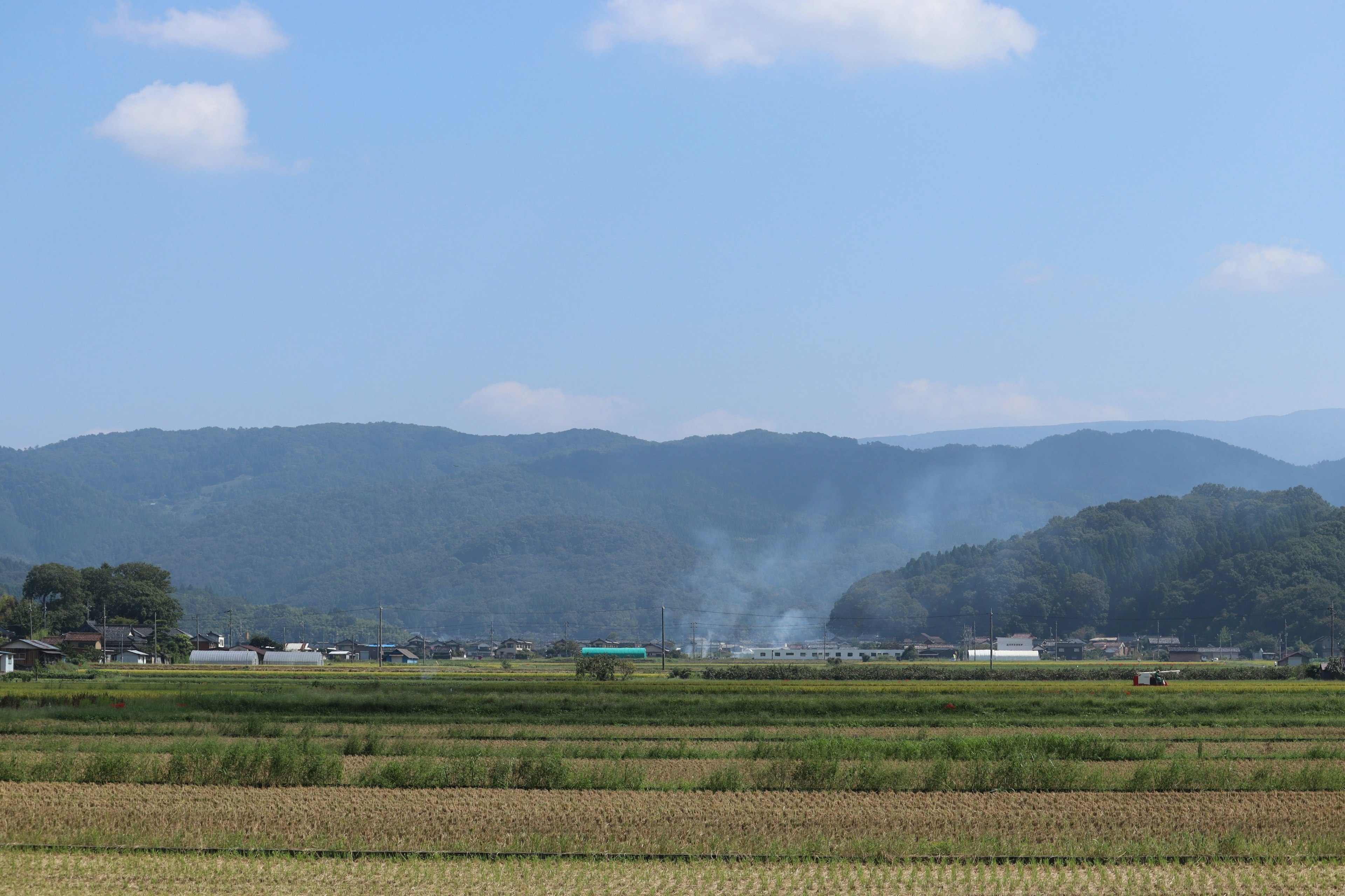 Rural landscape under blue sky with mountains in background and smoke rising