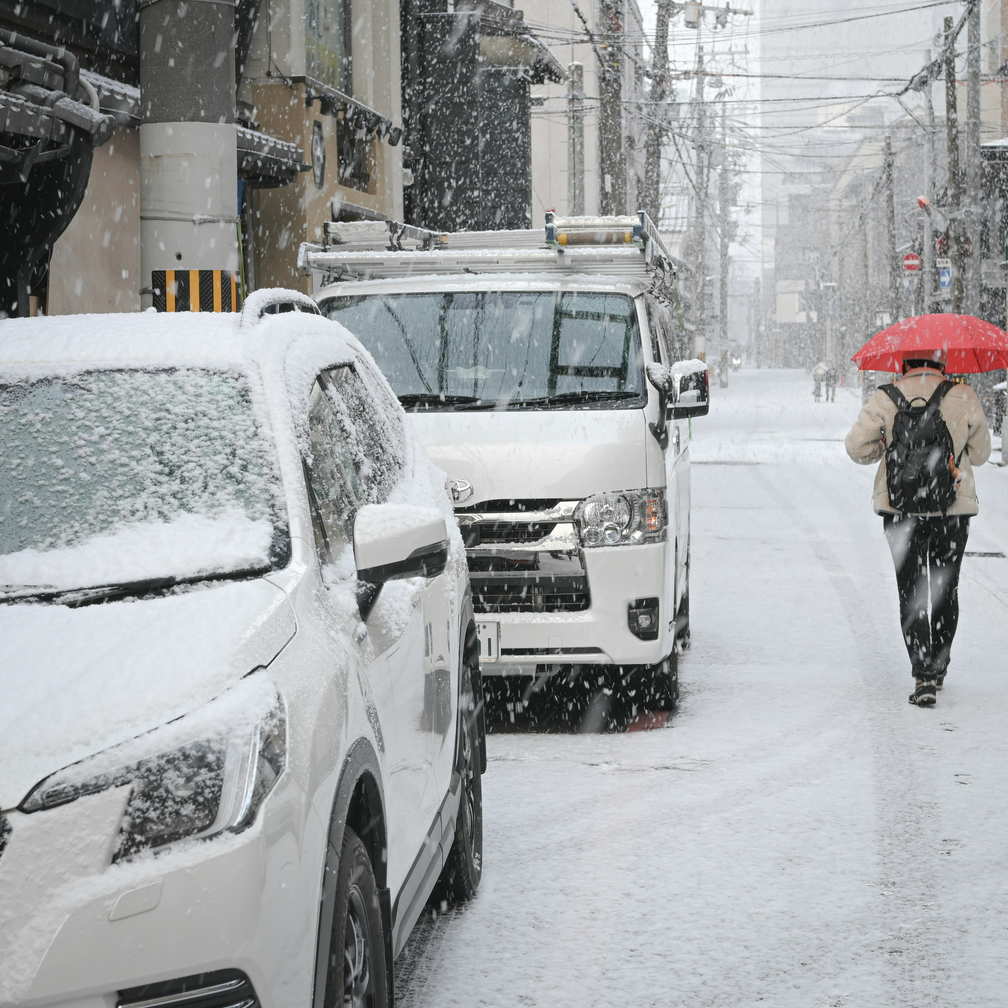 Eine Person, die mit einem roten Regenschirm auf einer verschneiten Straße geht, mit weißen Autos am Straßenrand