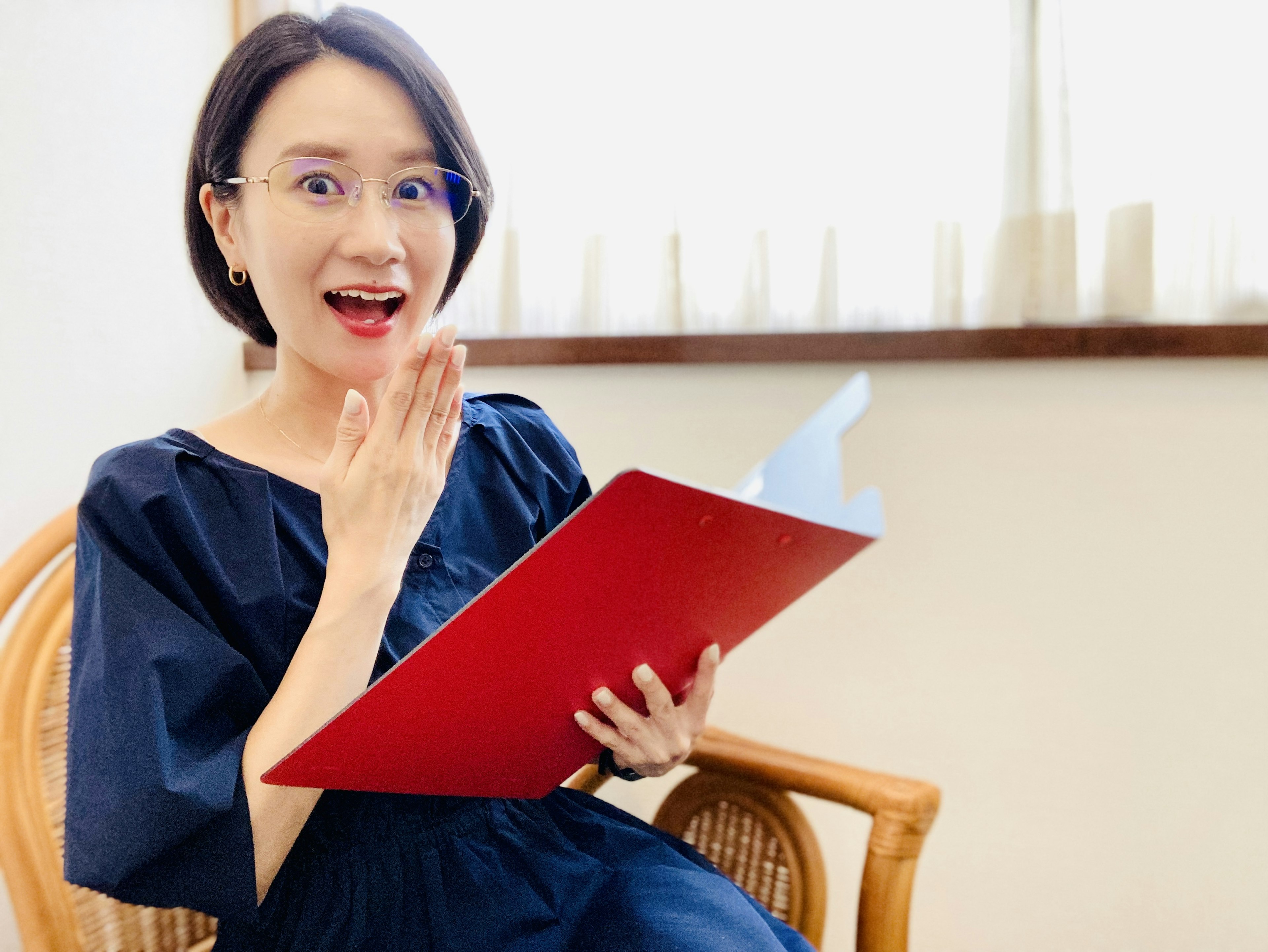 Une femme avec des lunettes tenant un dossier rouge et souriant dans un cadre intérieur