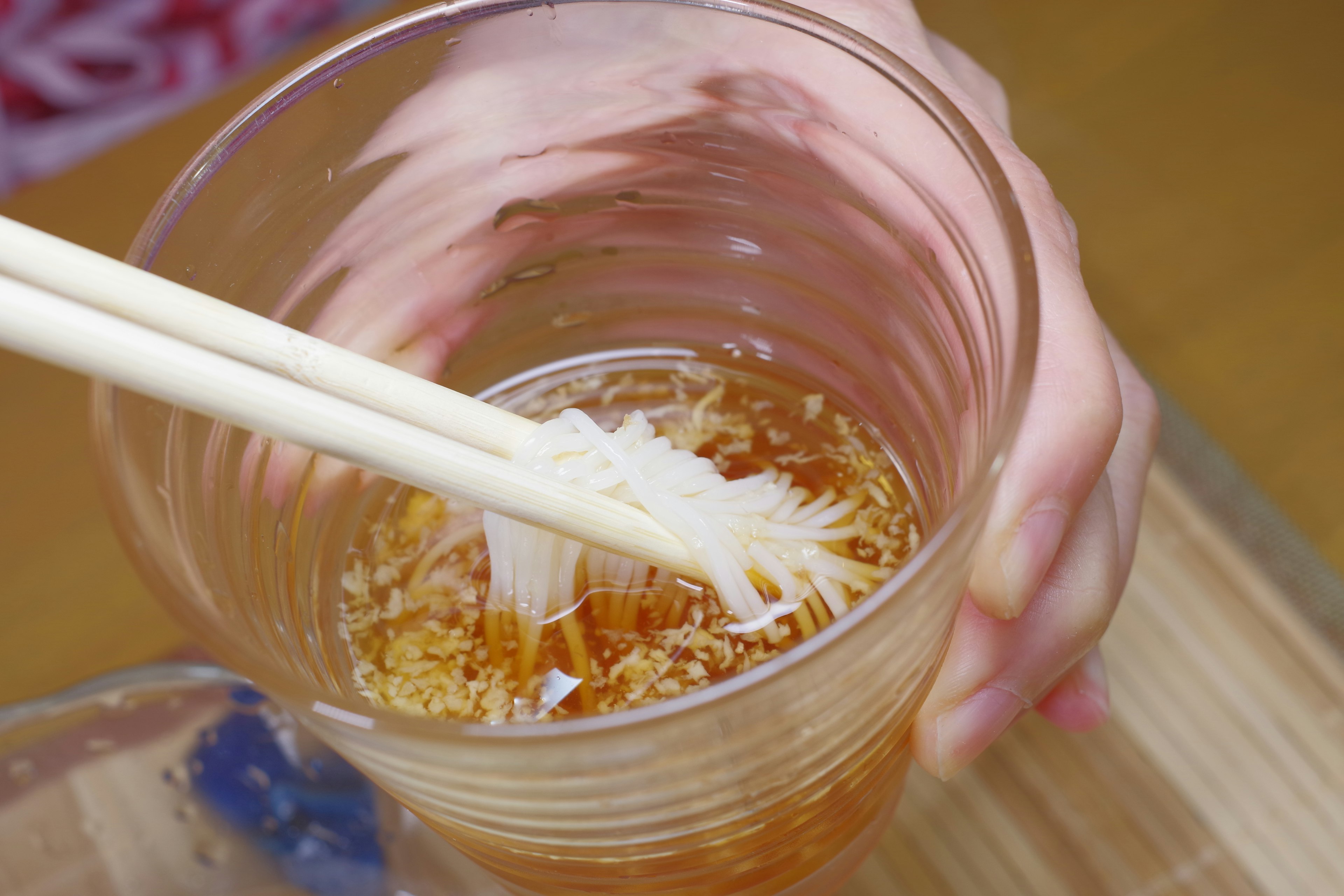 A hand lifting thin noodles with chopsticks from a transparent cup filled with liquid