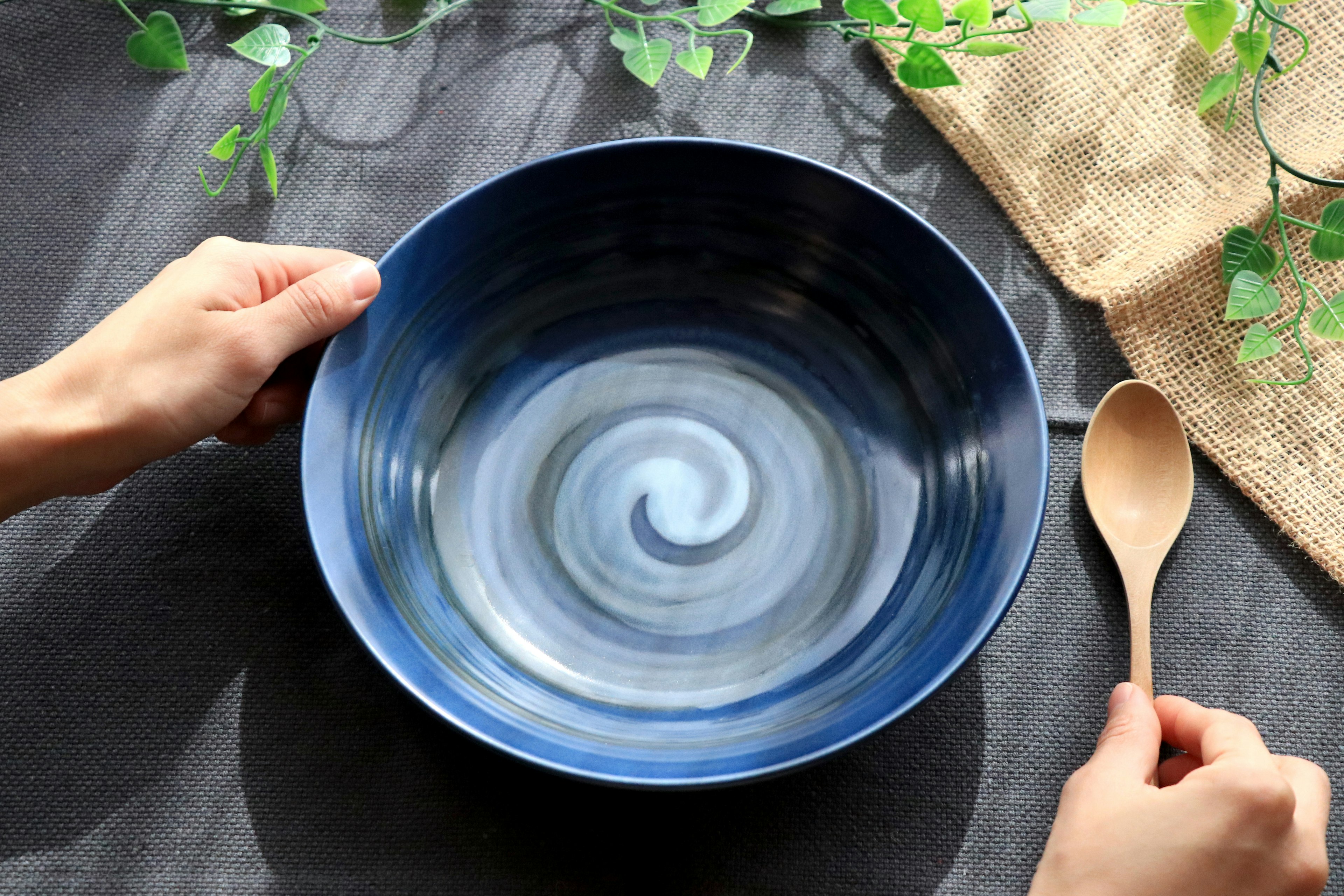 Hands holding a blue swirling bowl with a wooden spoon green leaves and burlap background