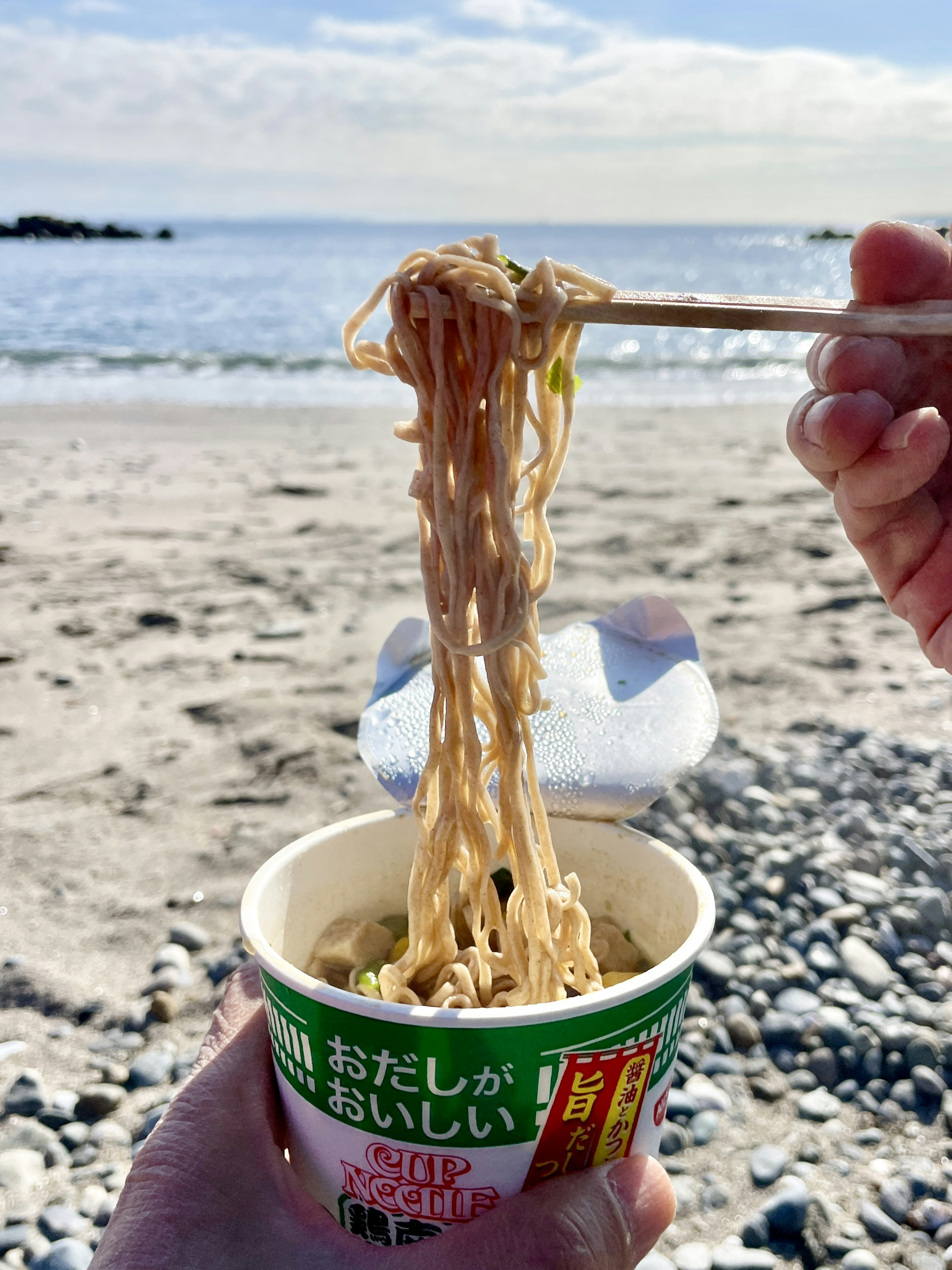 Eine Hand, die Nudeln aus einem Becher am Strand mit Meereshintergrund hält