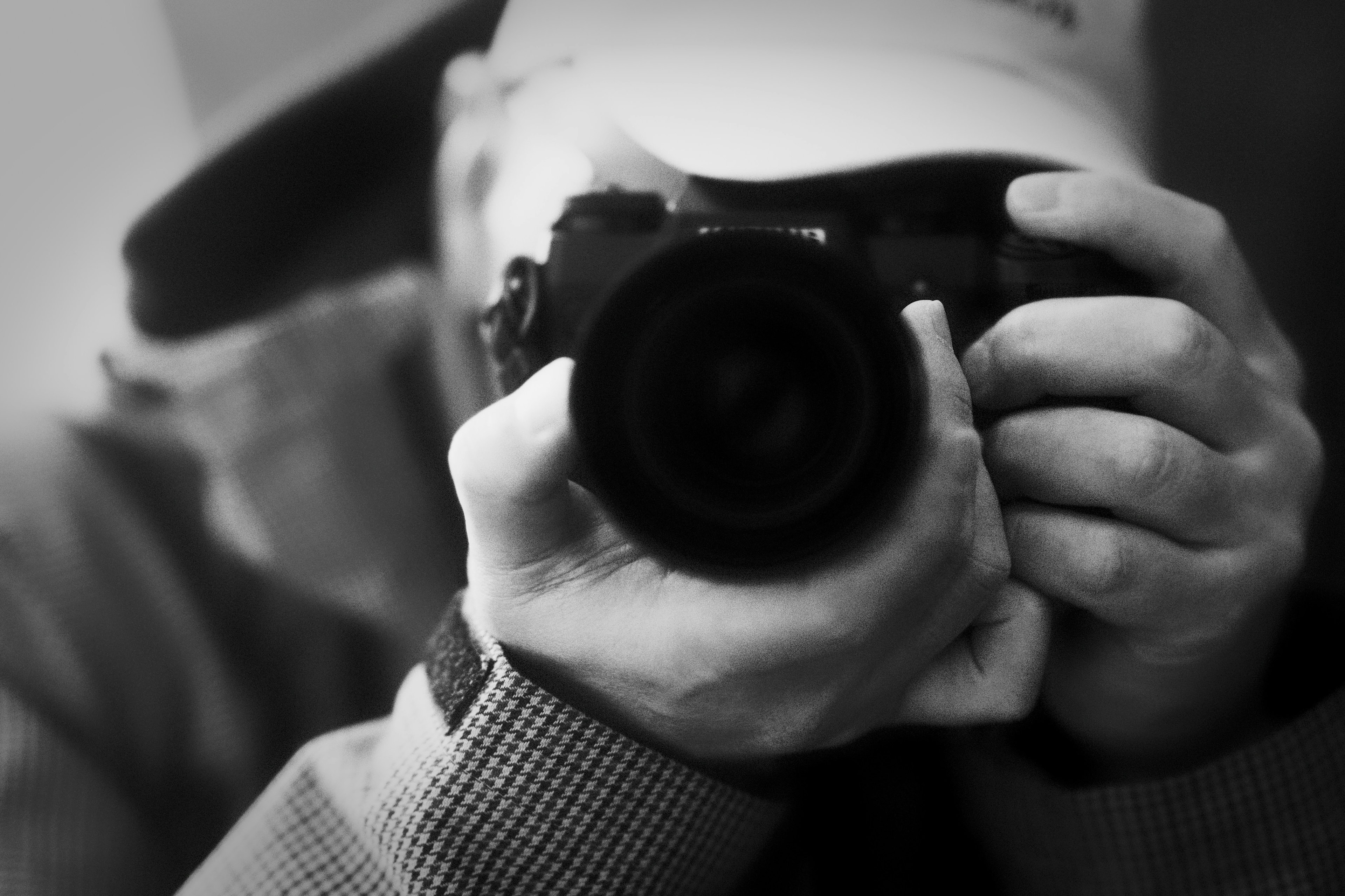 Black and white photo of a man holding a camera wearing a hat pointing the camera lens at the viewer