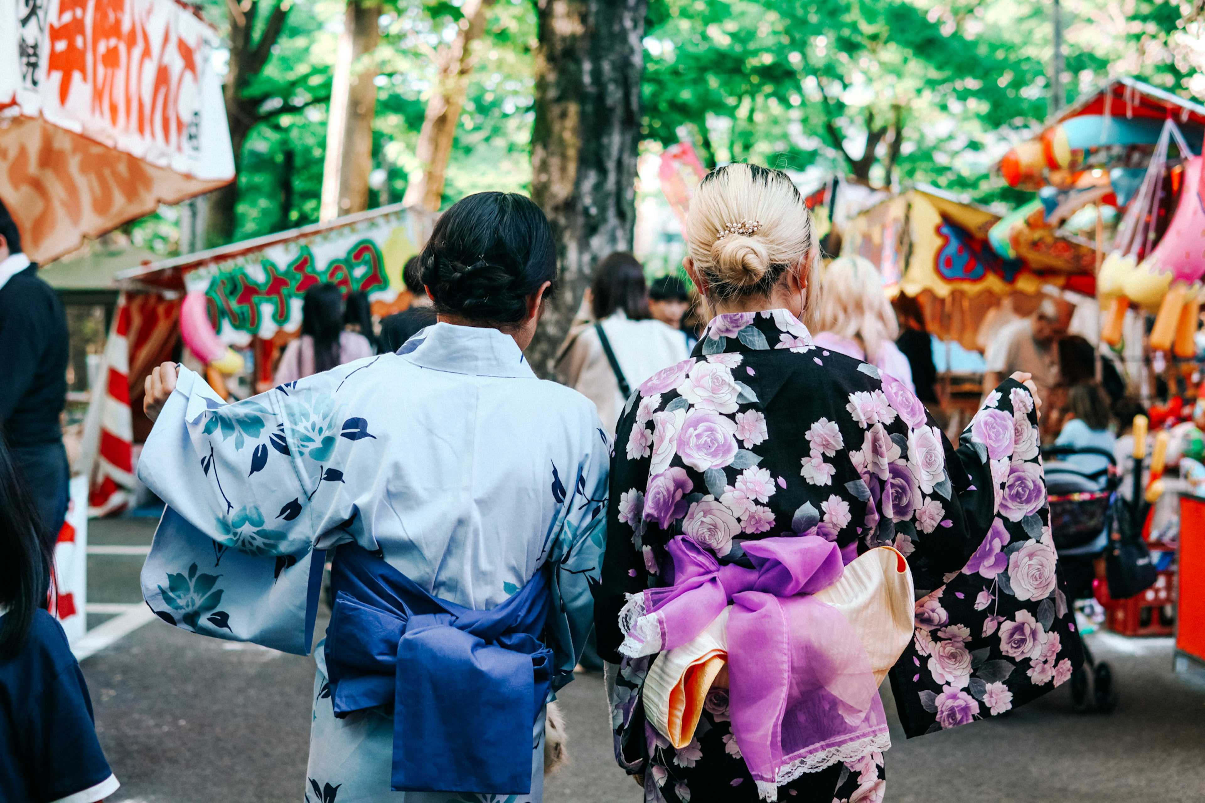 二人の女性が着物を着て祭りに向かう後ろ姿 色とりどりの装飾が施された屋台が背景にある