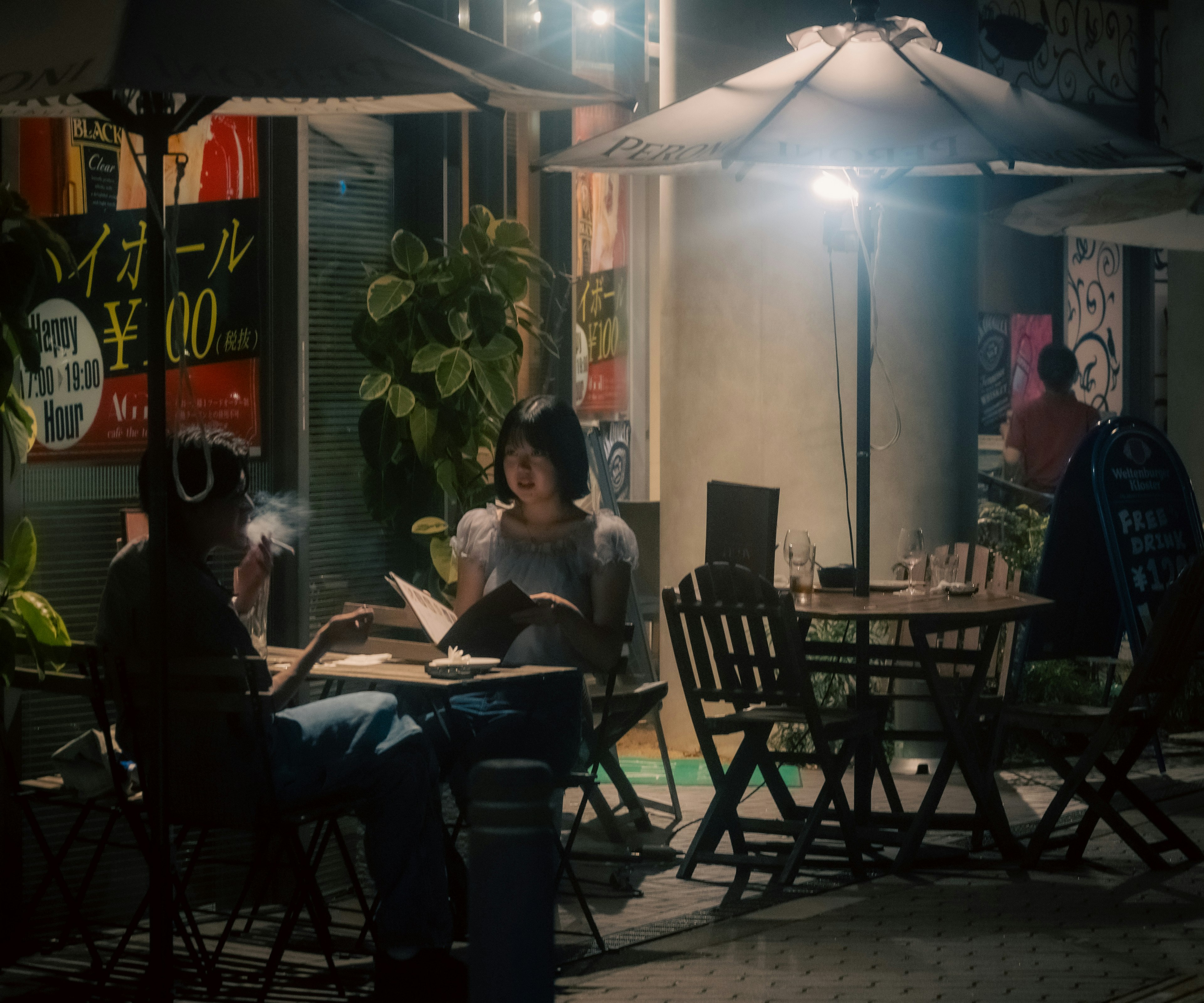 Silhouette einer Frau und eines Mannes, die in einem Nachtcafé unter einem Schirm mit Pflanzen um sich herum auf ein Menü schauen