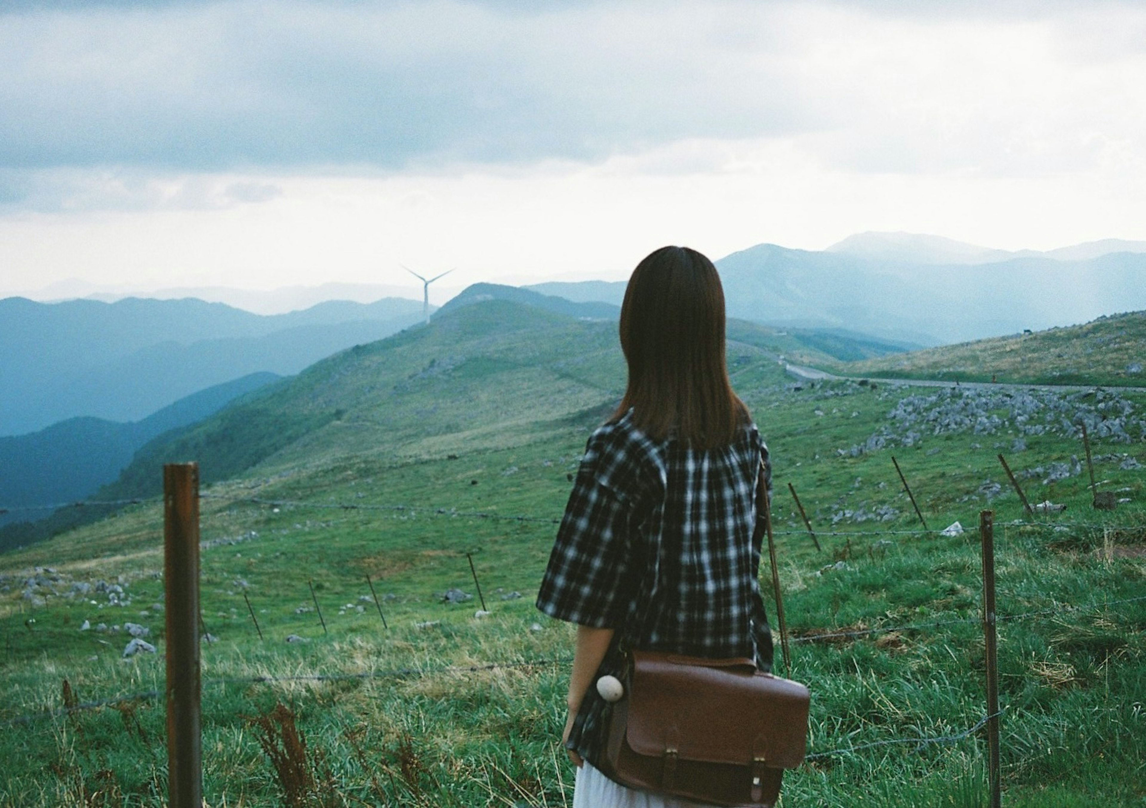 Girl in checkered shirt gazing at green hills with wind turbines