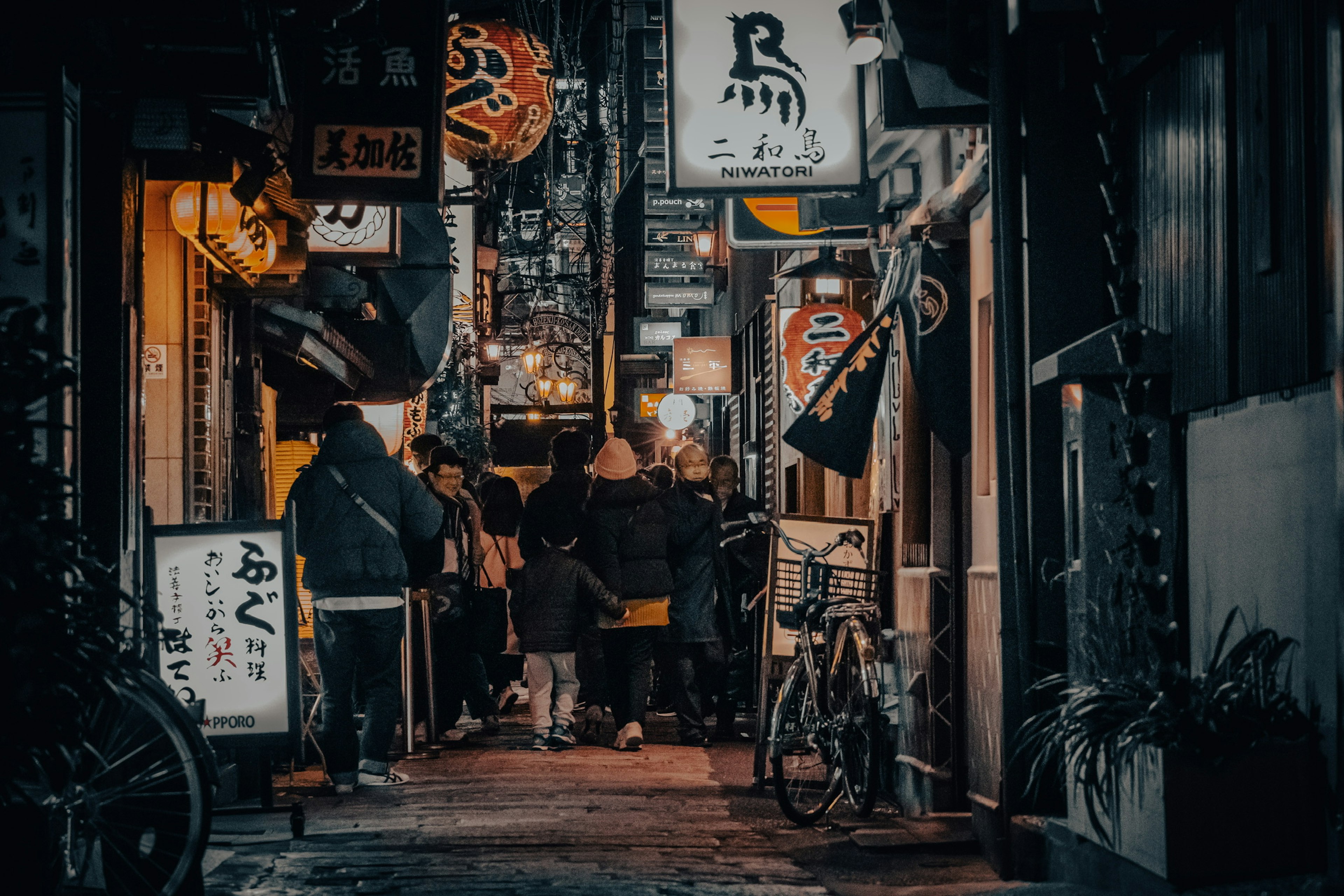 夜の賑やかな路地に人々が集まり、提灯や看板が灯る